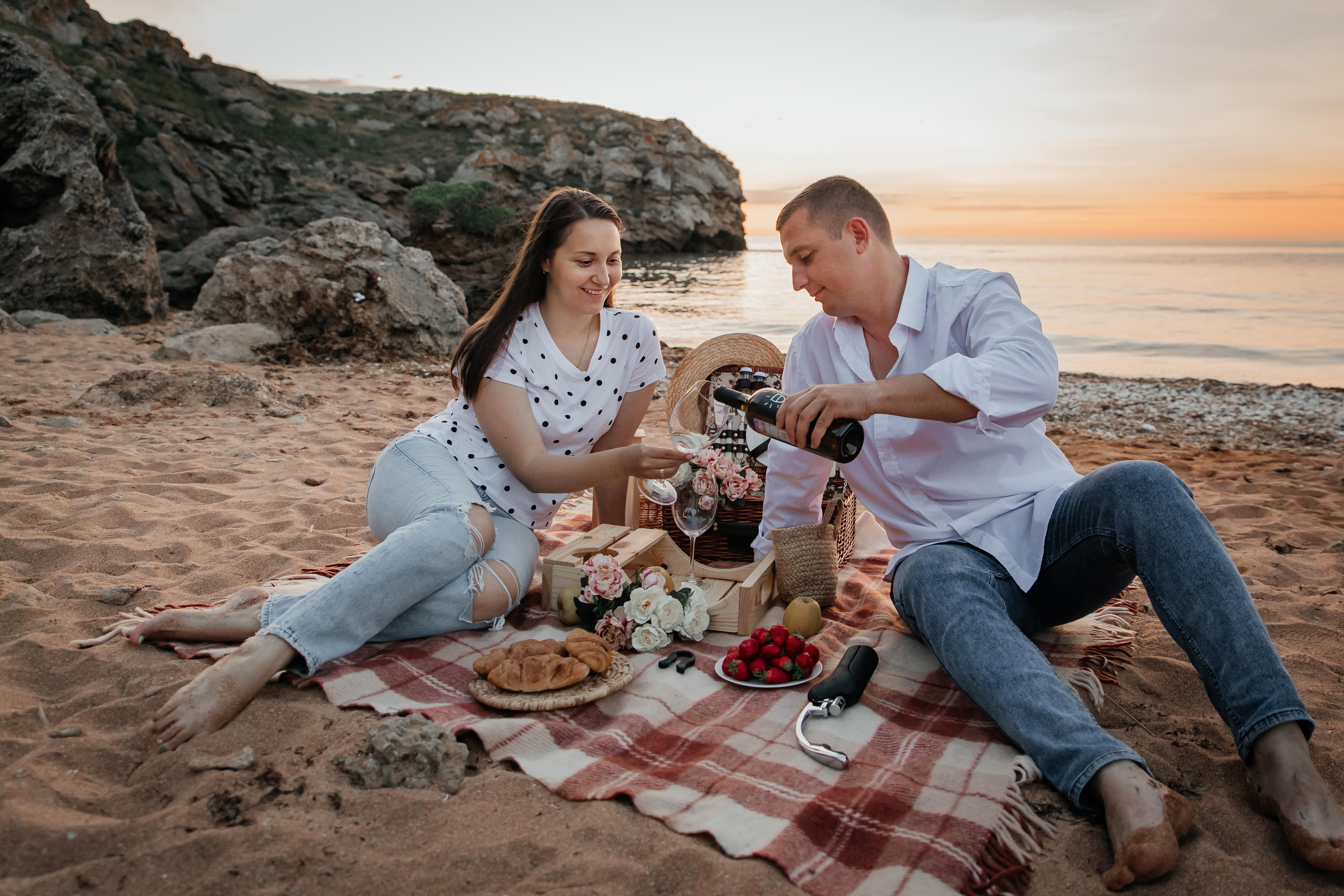 Пикник на пляже на закате , парень наливает вино в бокал девушке. 