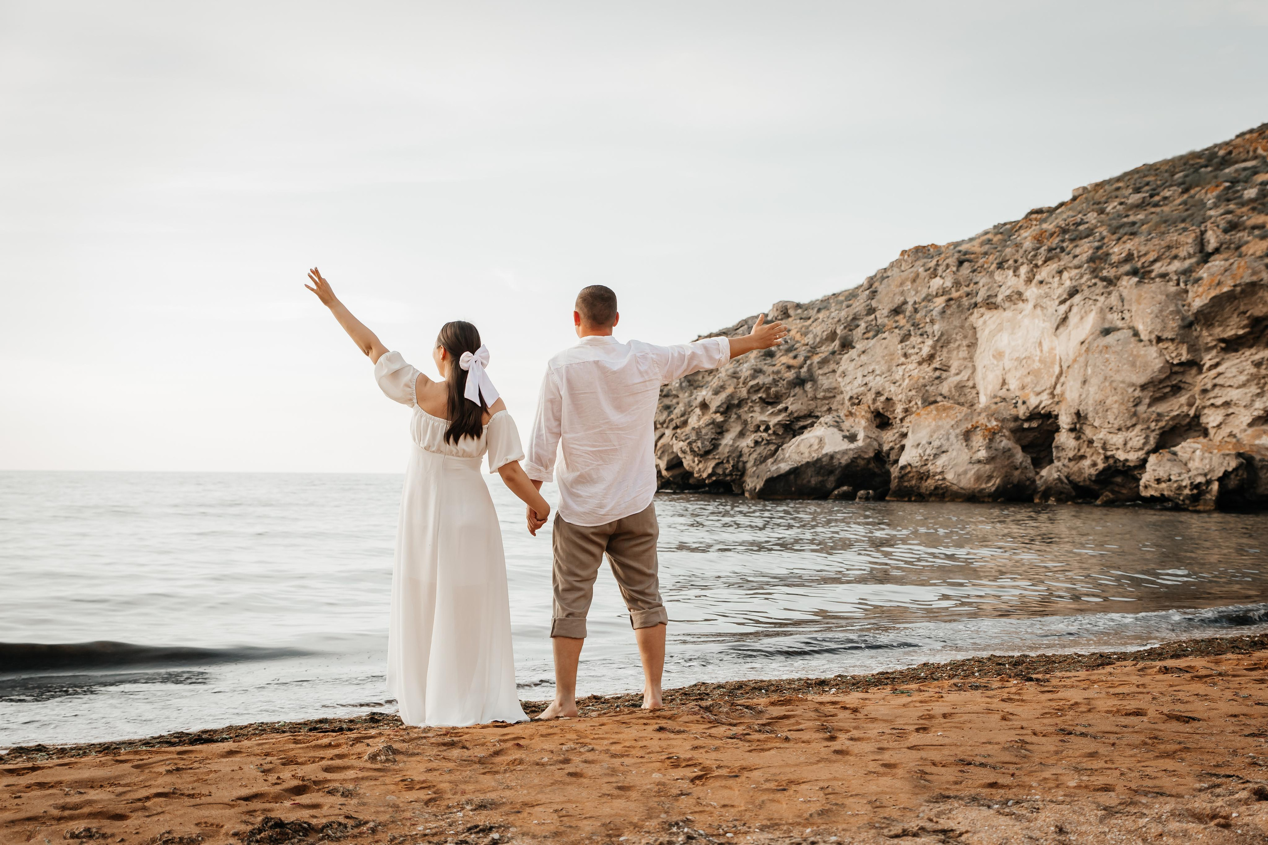 Девушка и парень стоят спиной на берегу моря на фоне скалы.
