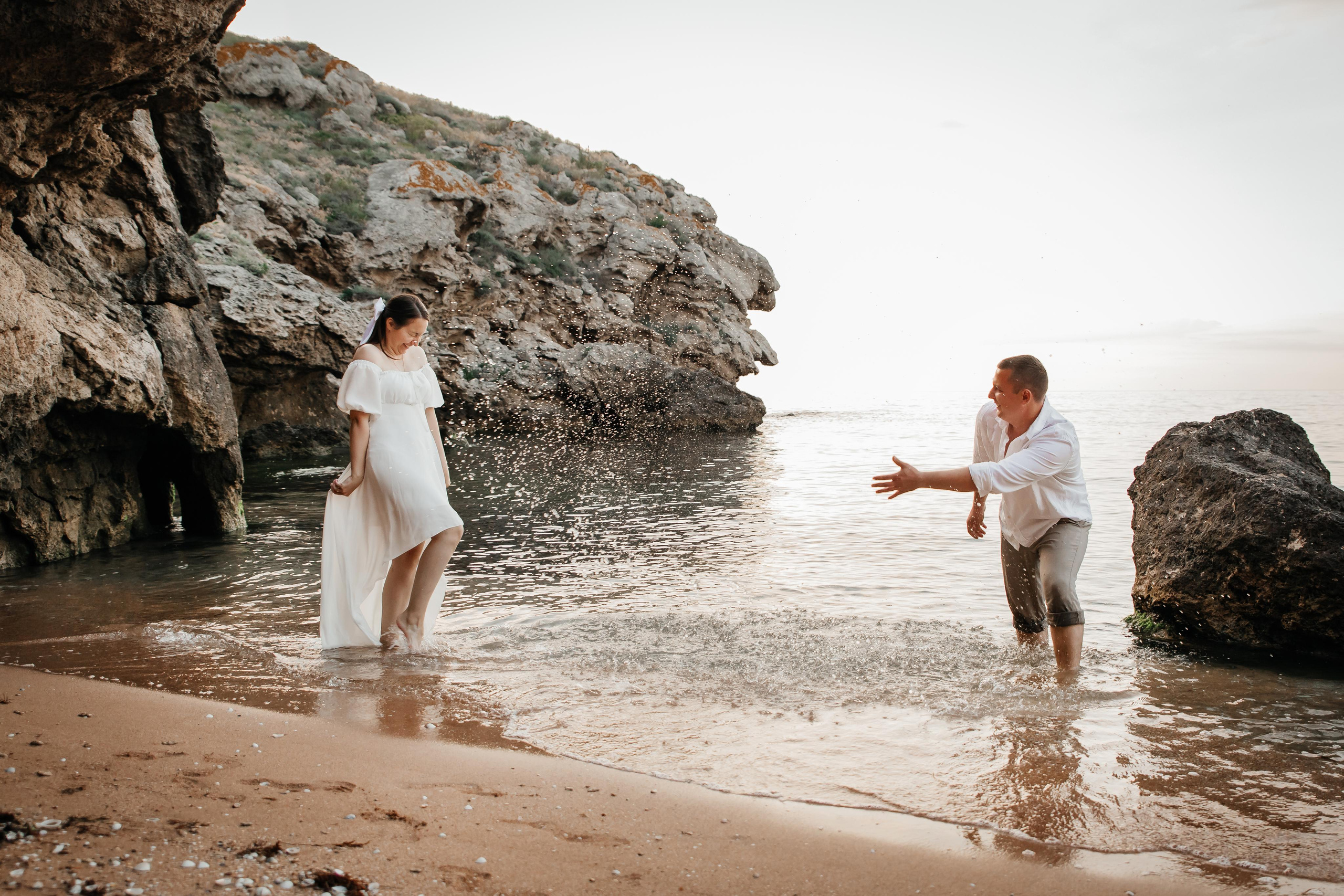 Парень брызгает на девушку водой на морском побережье на фоне скал. 