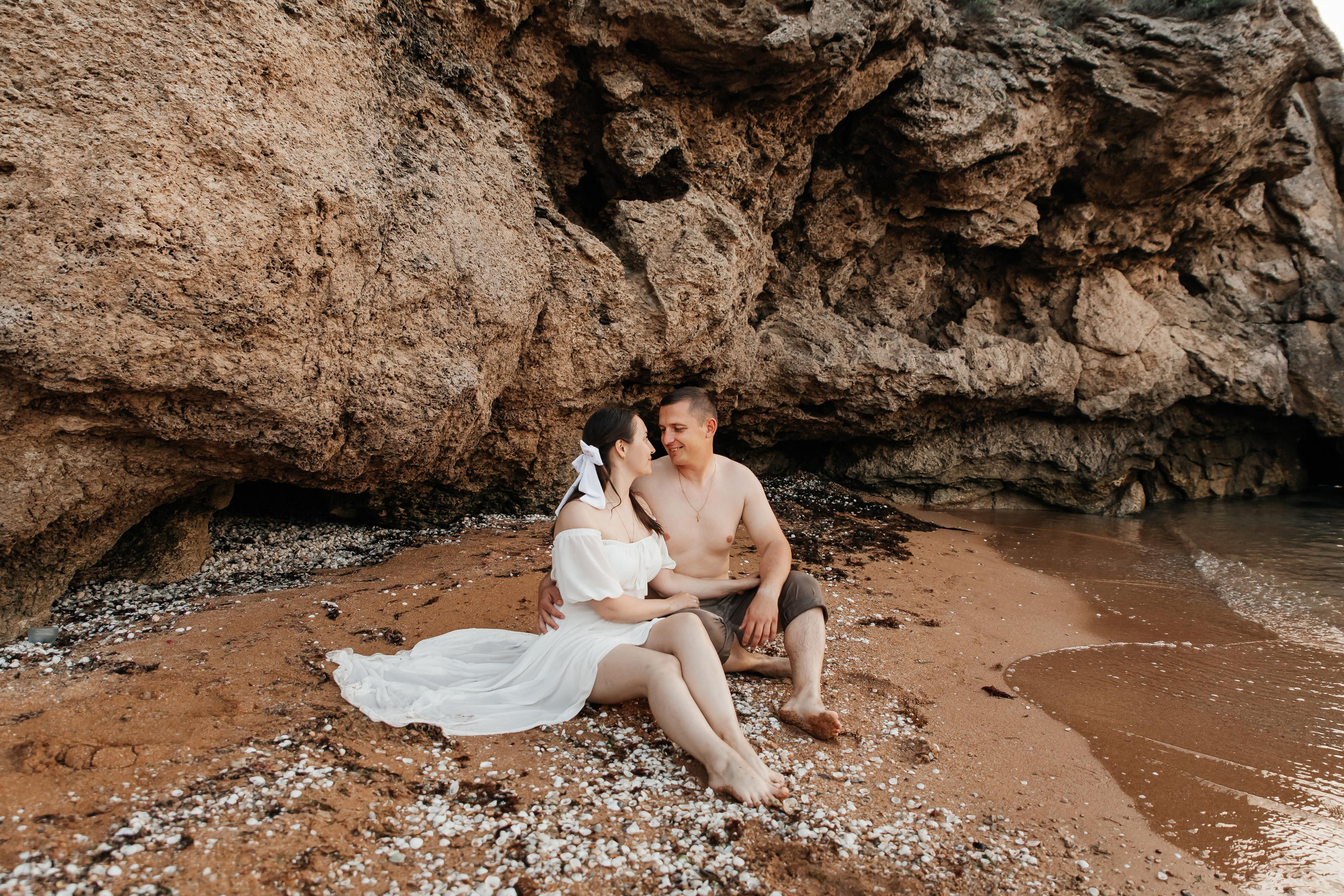 Пара сидит на песчаном пляже на берегу моря под скалами. 