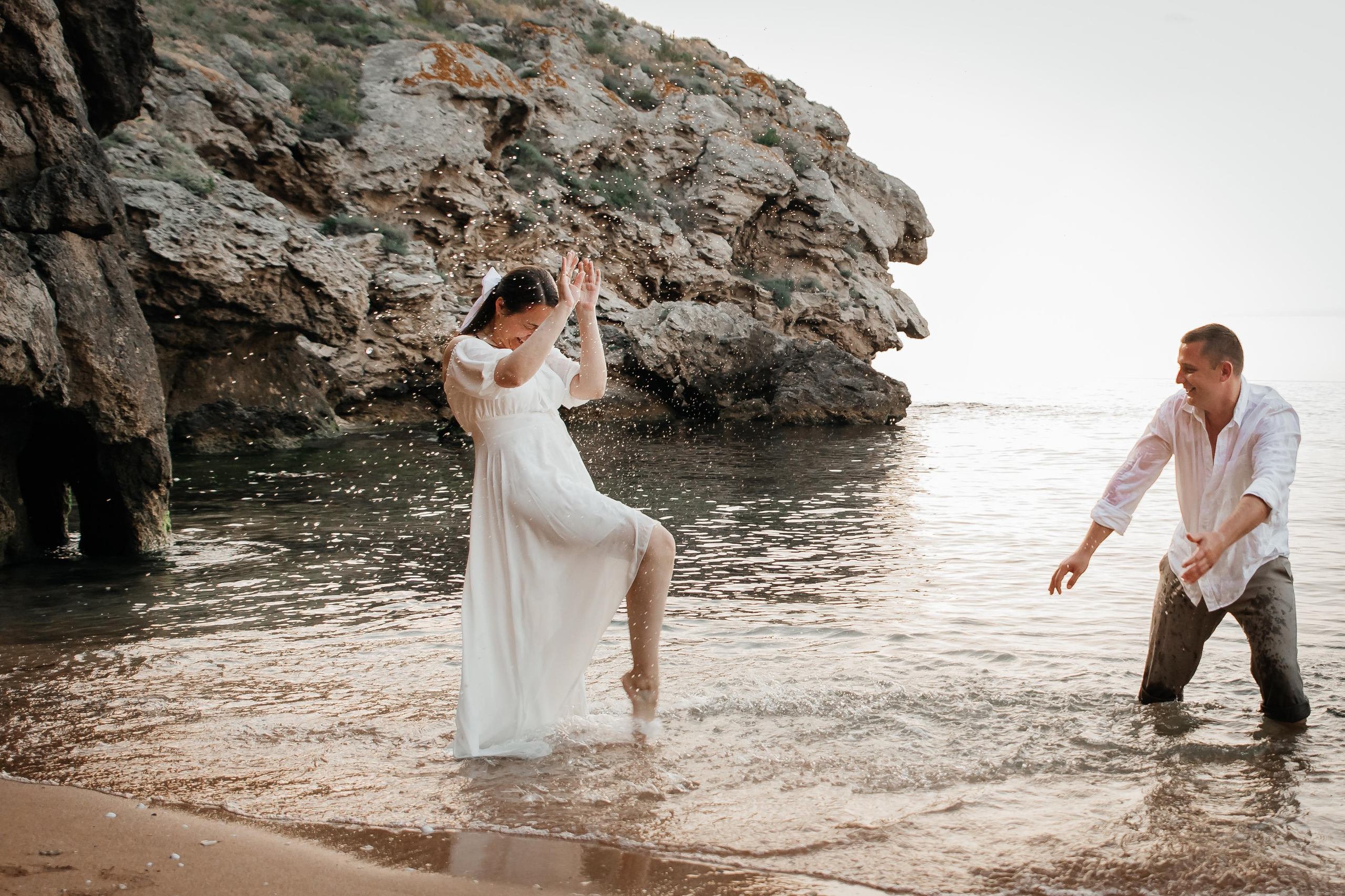 Девушка закрывается от брызг водой на морском побережье , парень брызгается на нее на фоне скал. 