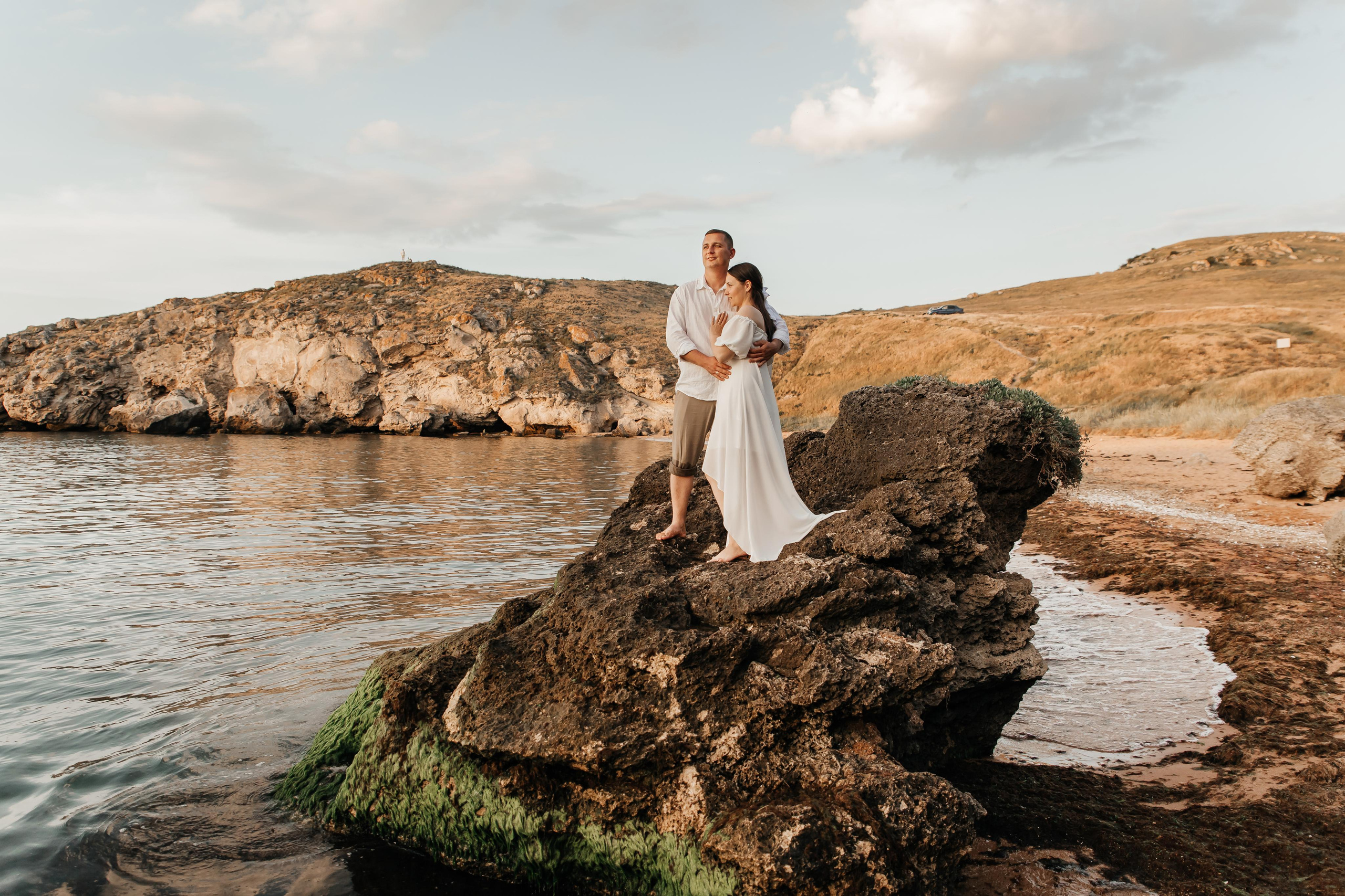 Девушка и парень в белом стоят на огромном камне на берегу моря и смотрят в даль. 