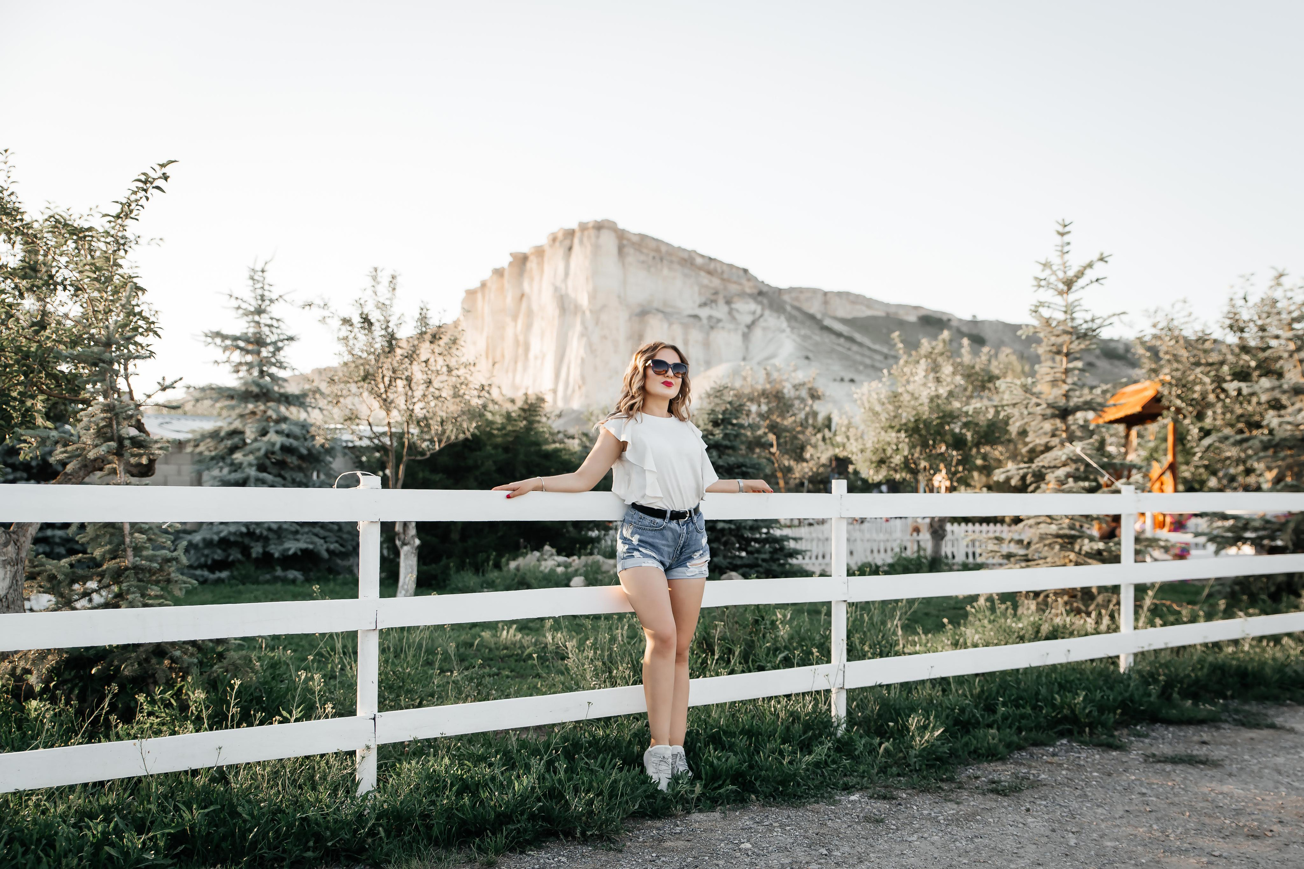 Девушка опирается на белое ограждение и смотрит вдаль на фоне скалы.