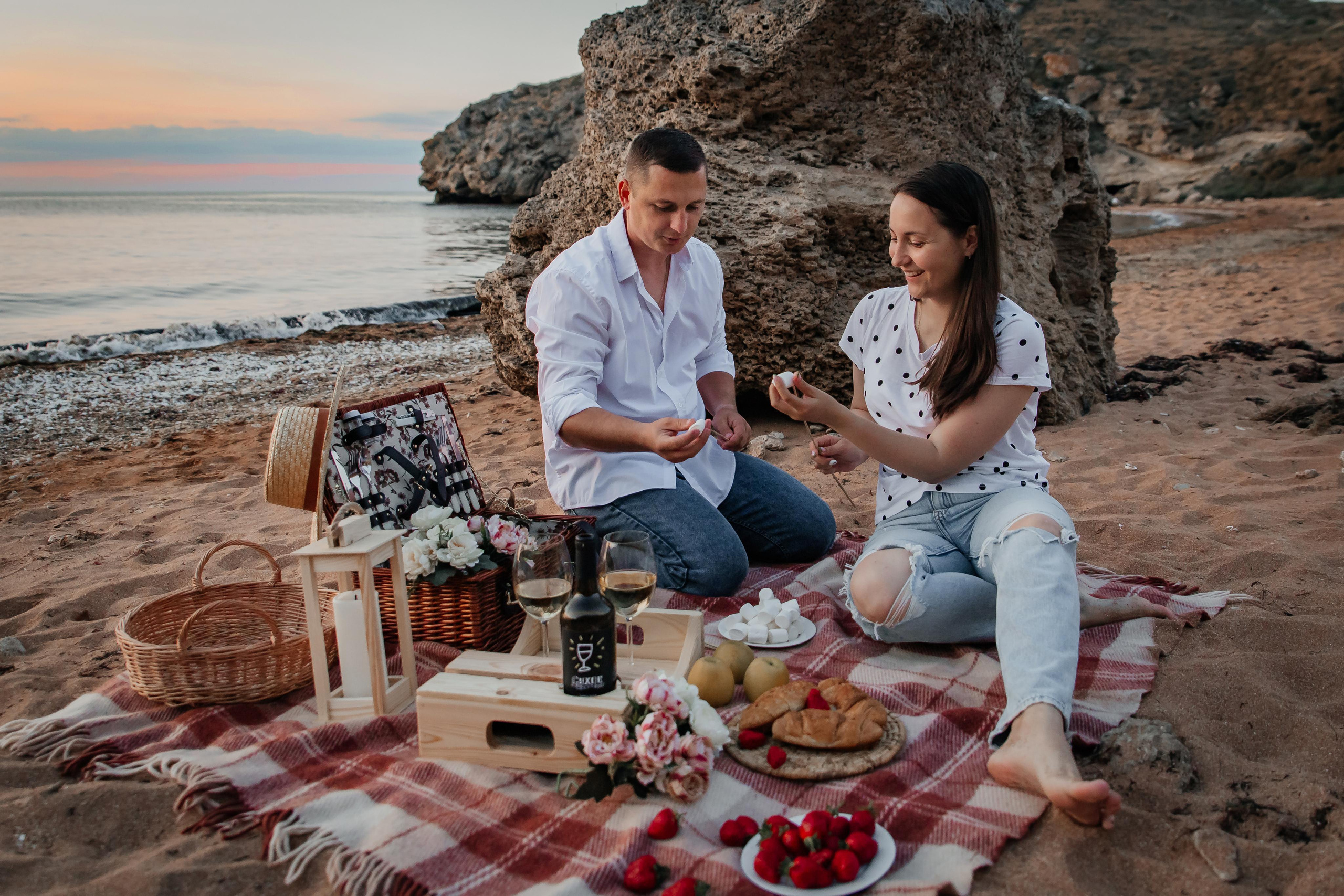 Пикник на морском побережье, девушка и парень сидят на пледе и одевают зефир на шпажки. 