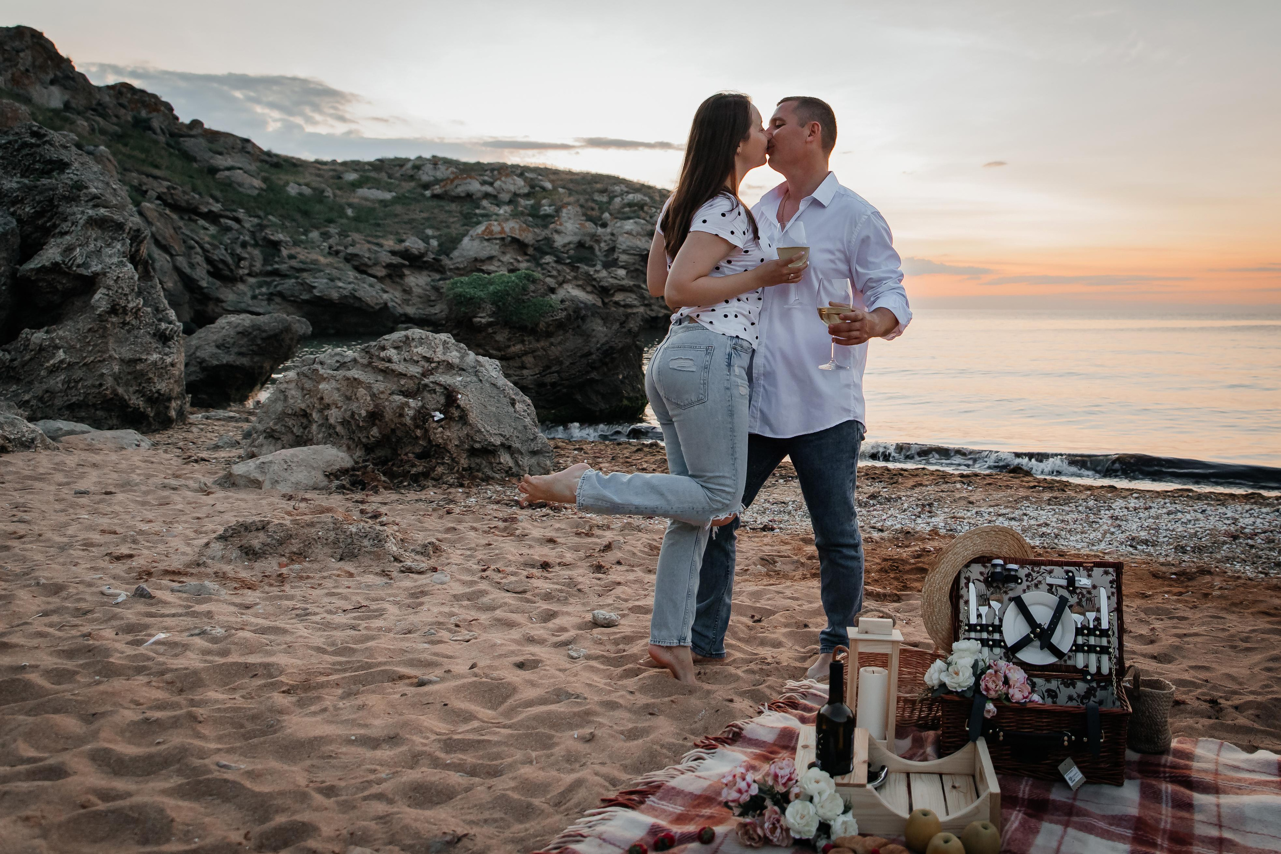 Пара стоит на песчаном пляже у них пикник, он держат бокалы с вином в руках, девушка целует парня.