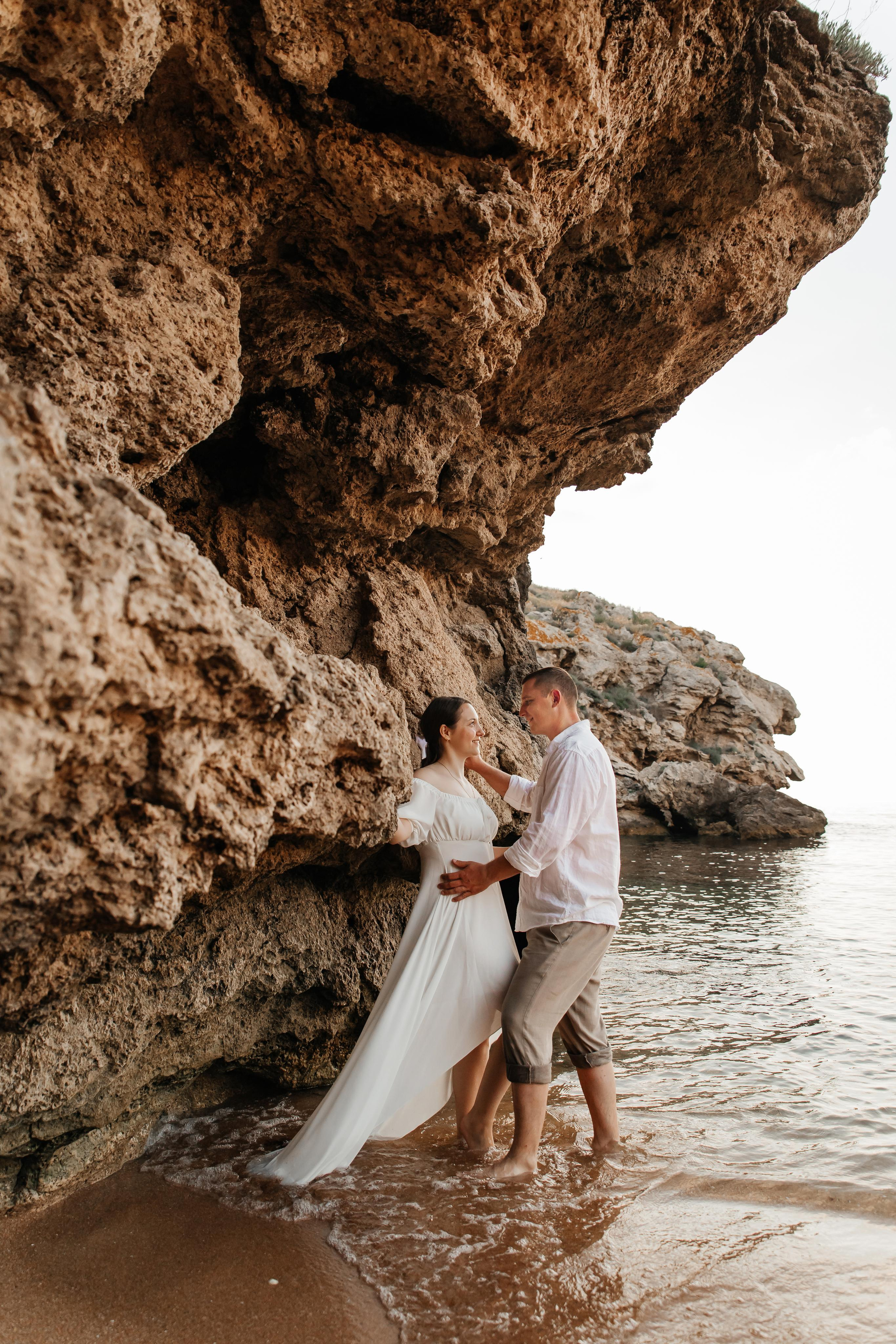 Пара в белом обнимается стоя в морском прибое под скалой. 