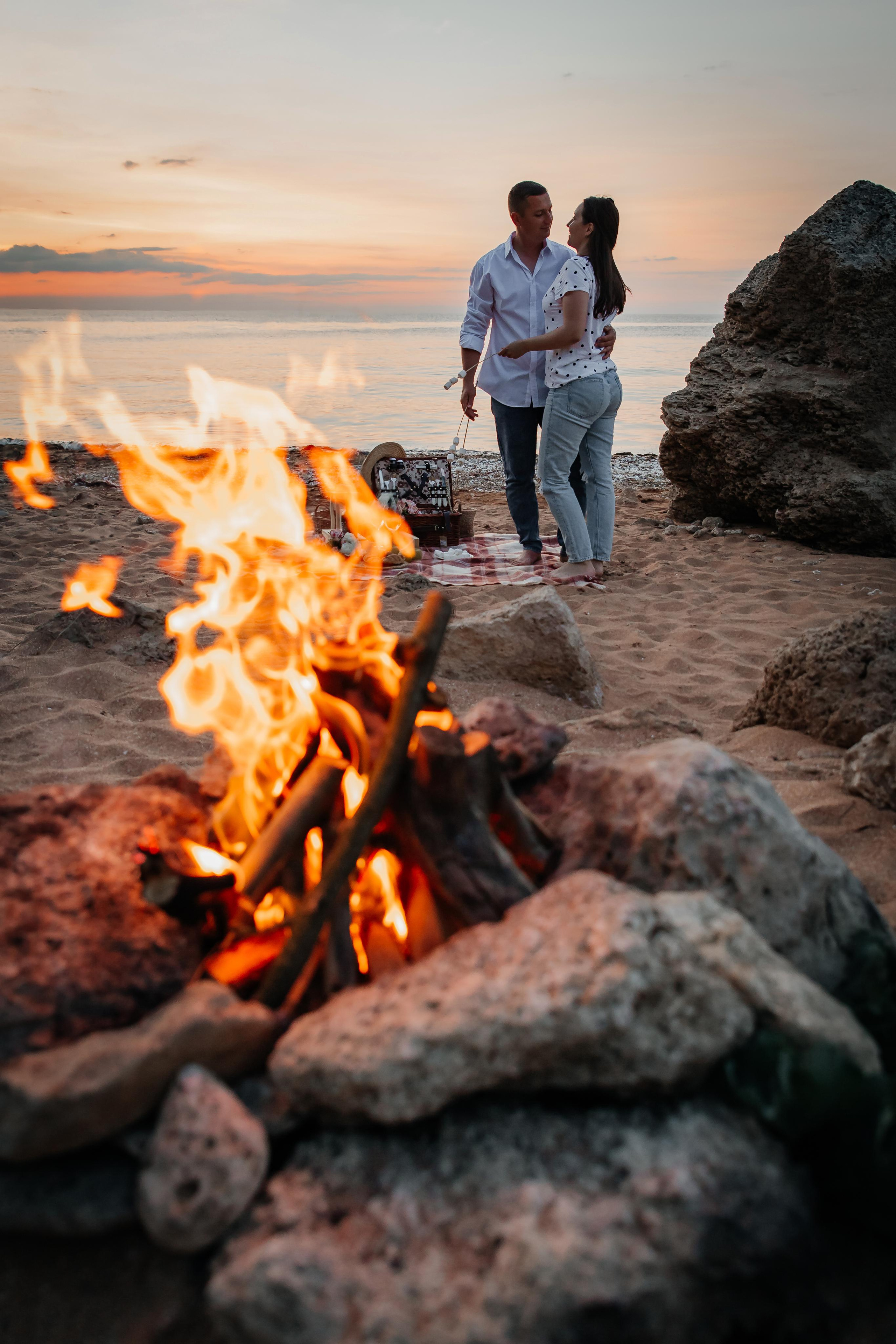 На переднем плане костер на песчаном пляже на берегу моря , вдалеке стоит влюбленная пара на закате. 