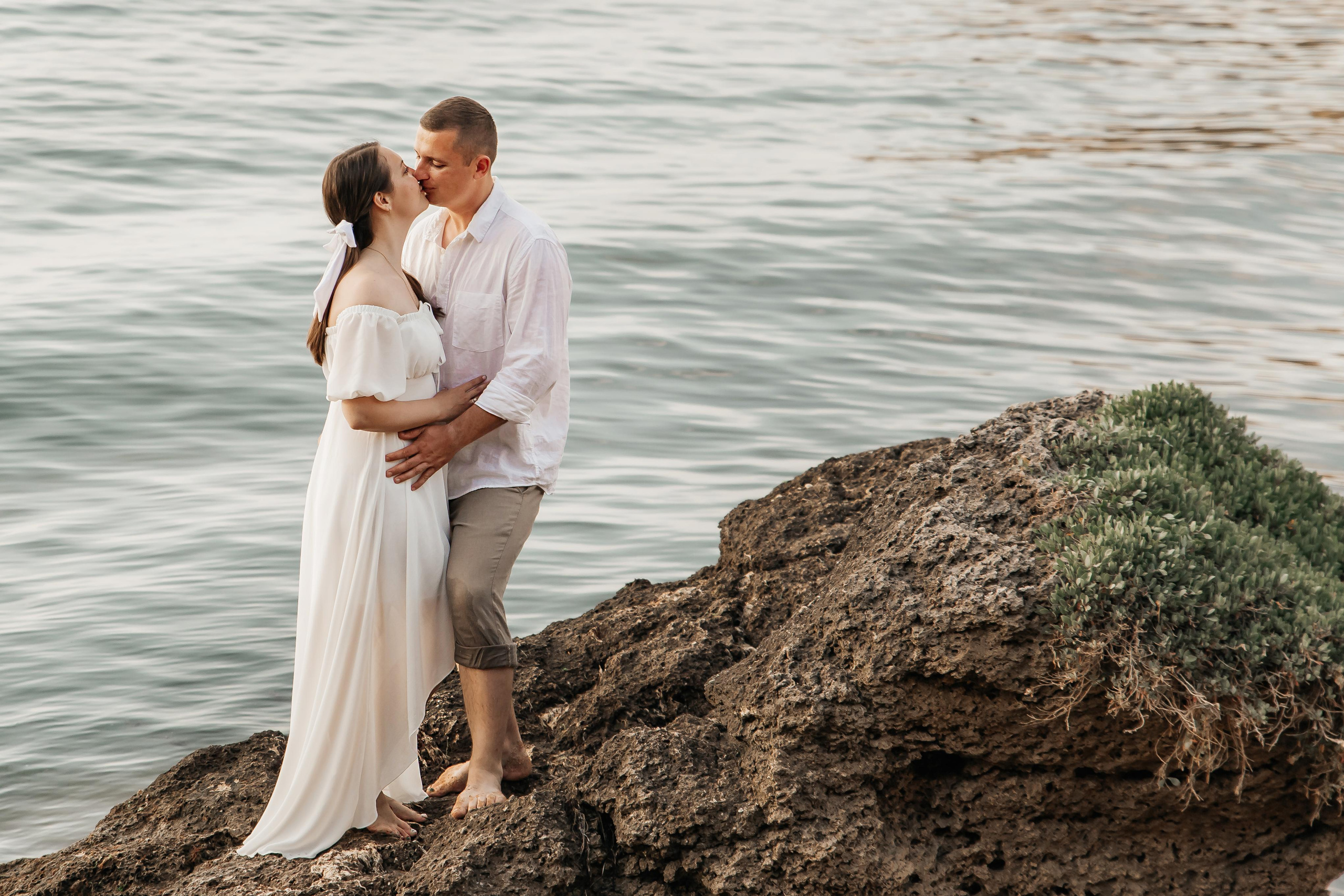 Целующаяся пара в белом на огромном камне на фоне моря крупным планом.