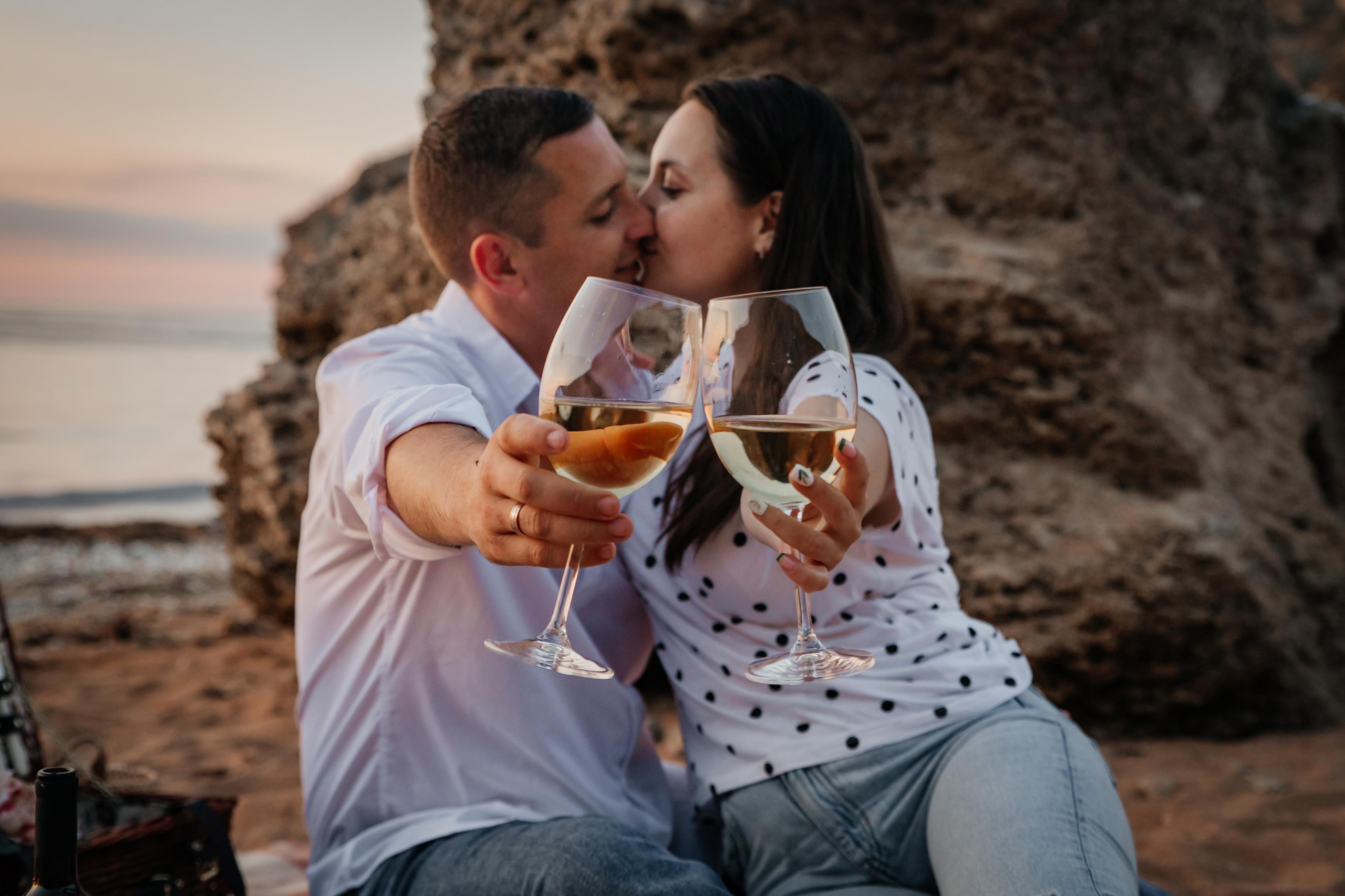 На переднем плане бокалы с вином их держит влюбленная целующаяся пара на закате на пляже . 