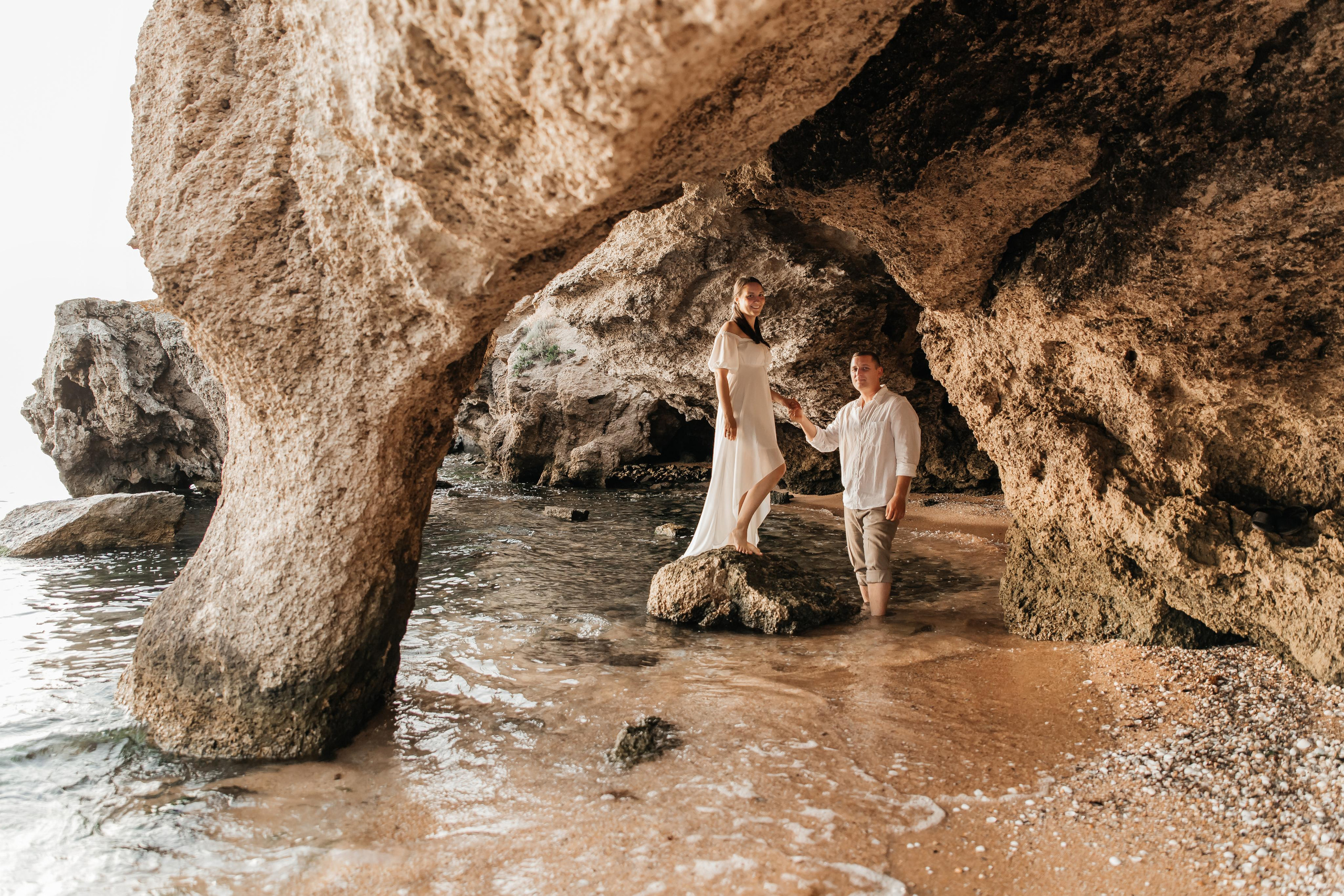 Девушка стоит на камне в море , парень держит ее за руку , над ними нависает скала.