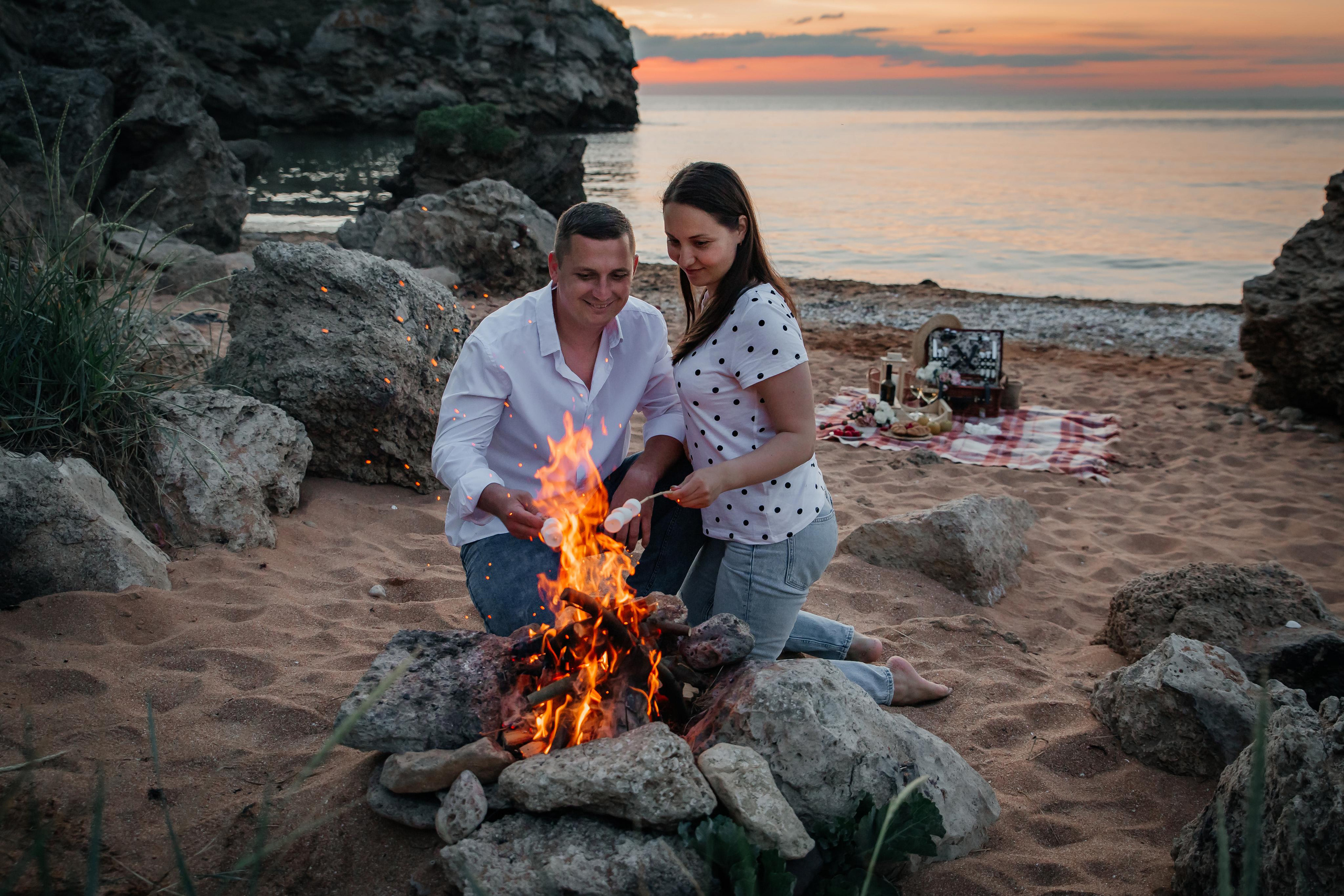 Пара сидит у костра на морском берегу на закате и жарит зефир на костре. 