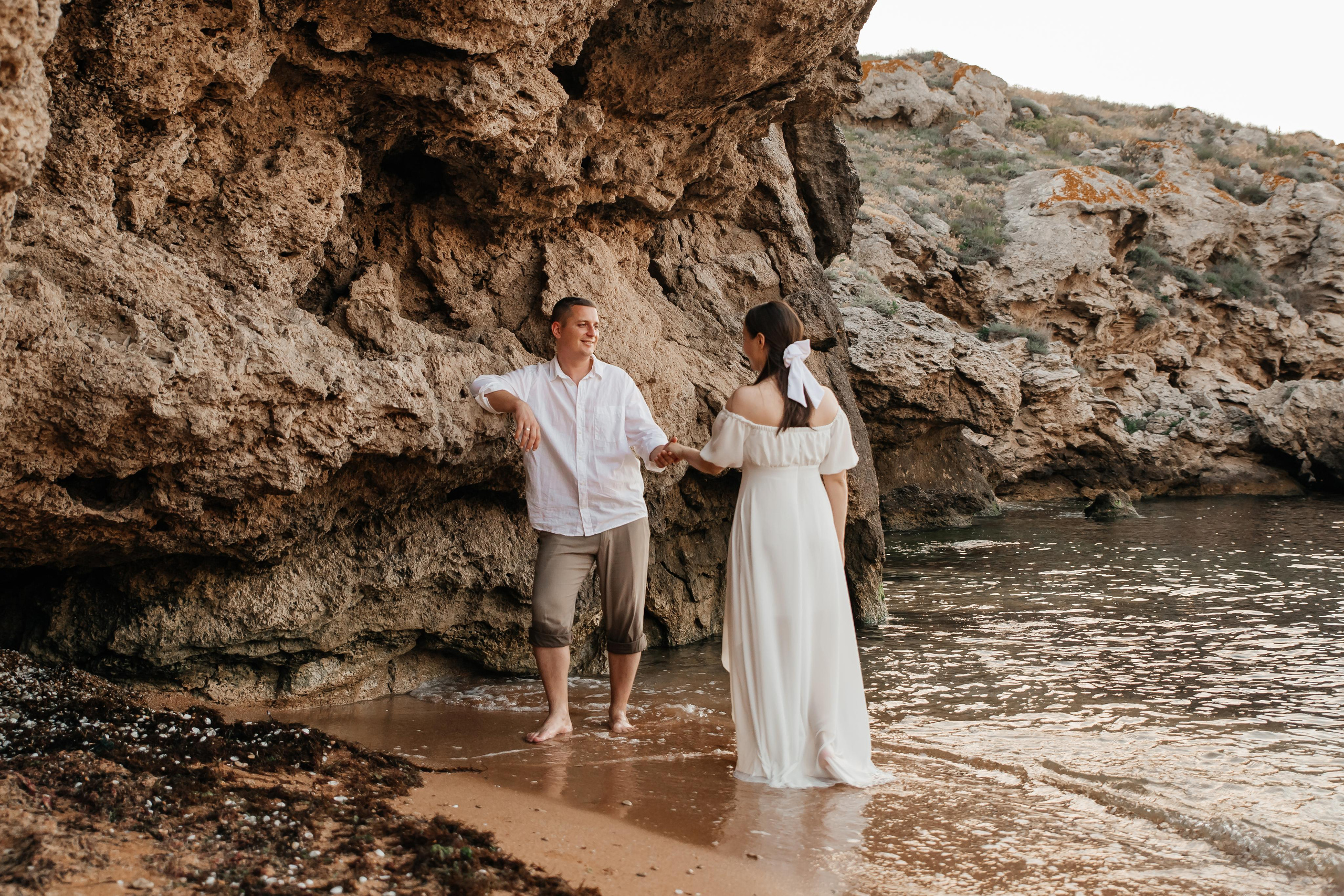 Под скалой стоит парень , к нему подходит девушка в белом на морском побережье. 