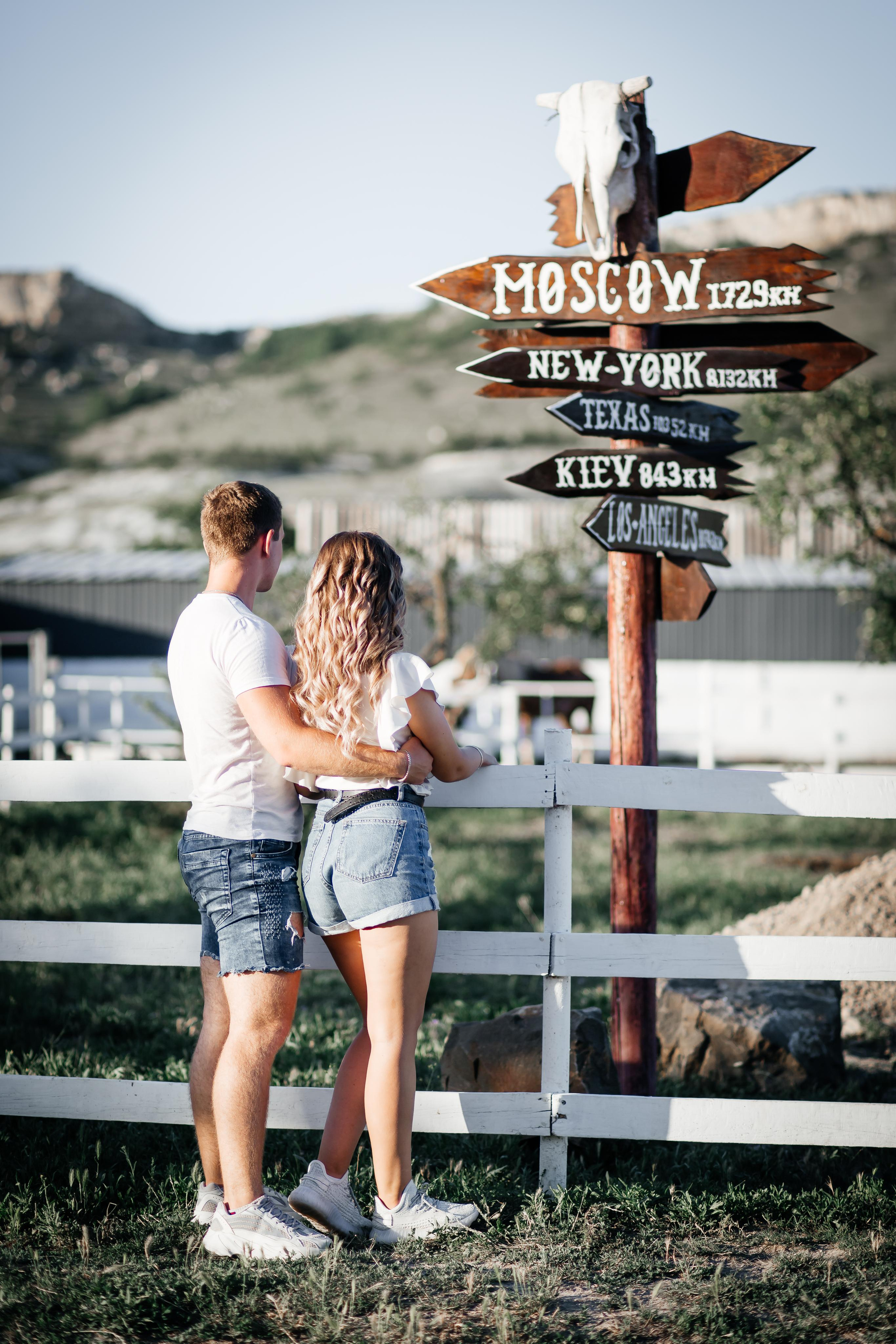 Пара стоит спиной обнявшись на фоне красивого указателя дорог.