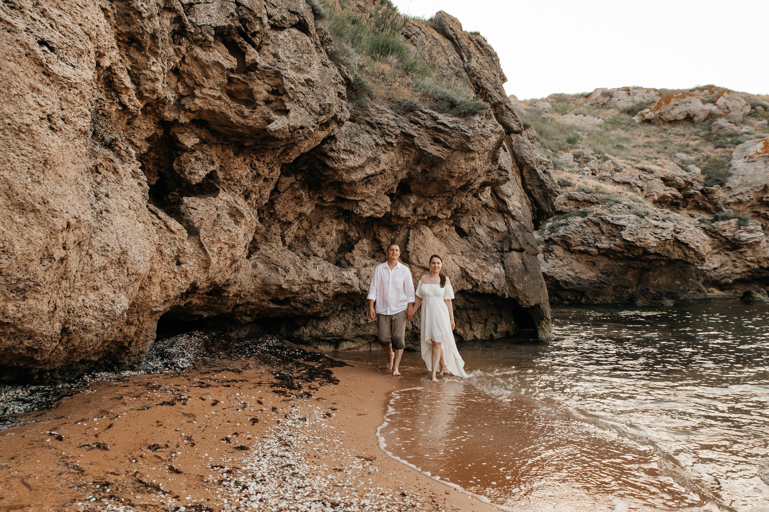 Пара идет по песчаному пляжу в морском прибое на фоне скал. 