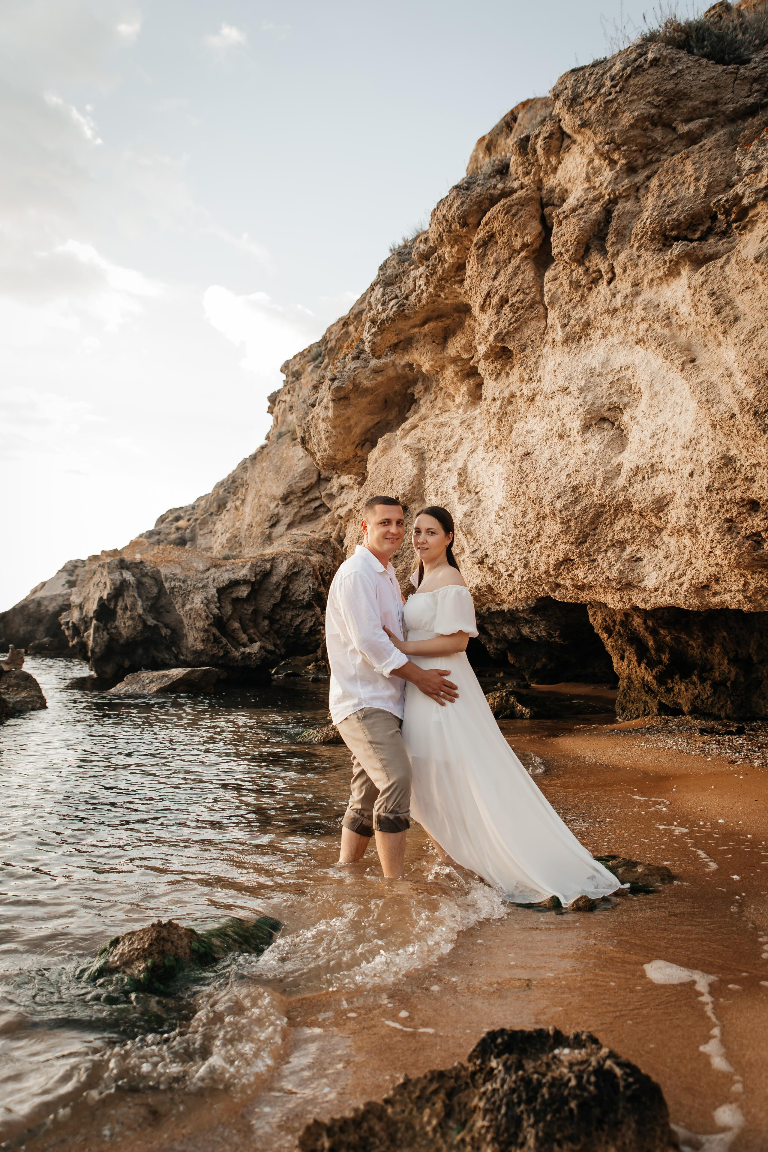 Девушка и парень в белых одеждах обнимаются на берегу моря на фоне больших скал. 