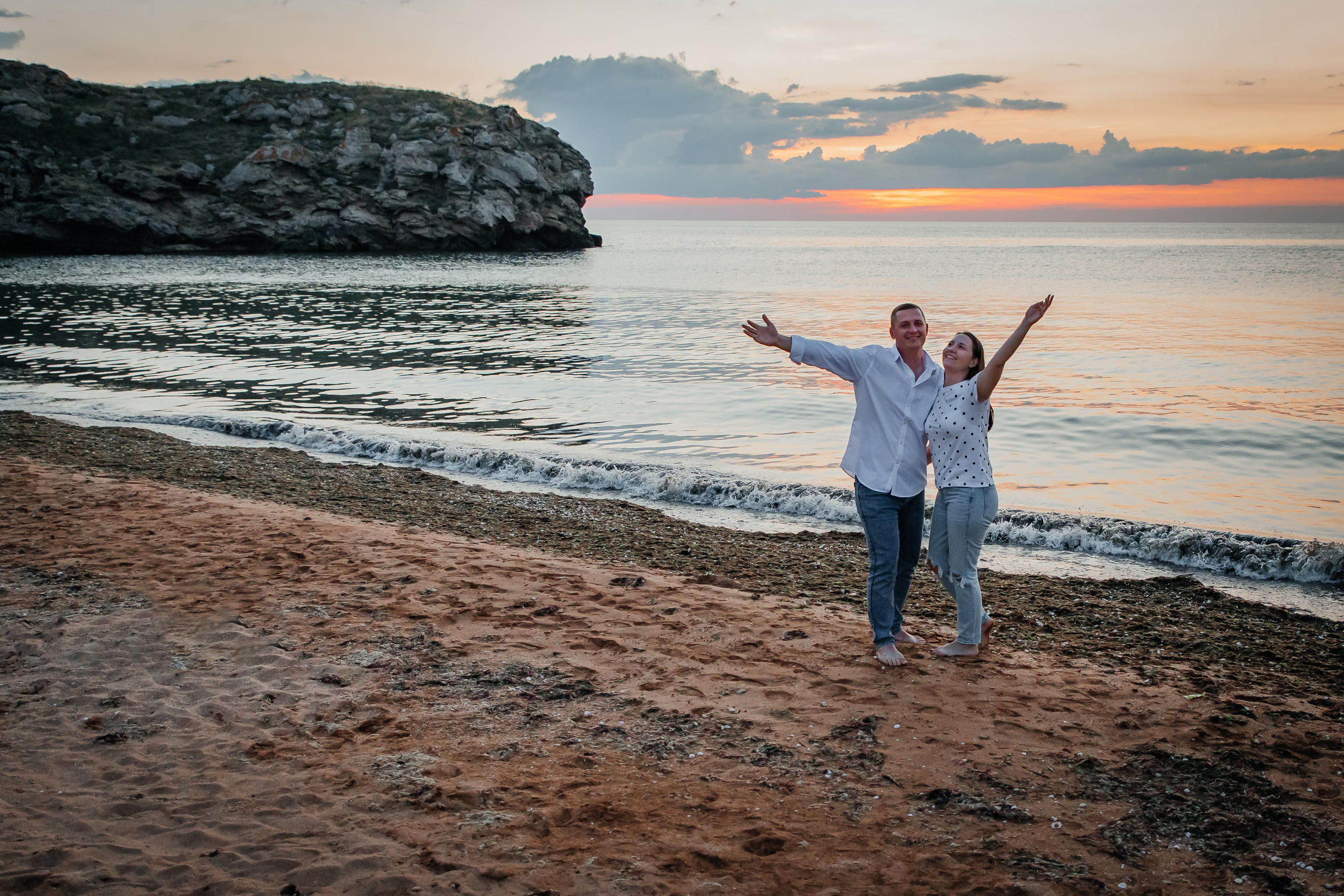 На морском берегу на закате стоит обнявшись влюбленная пара , они улыбаются и подняли руки верх. 