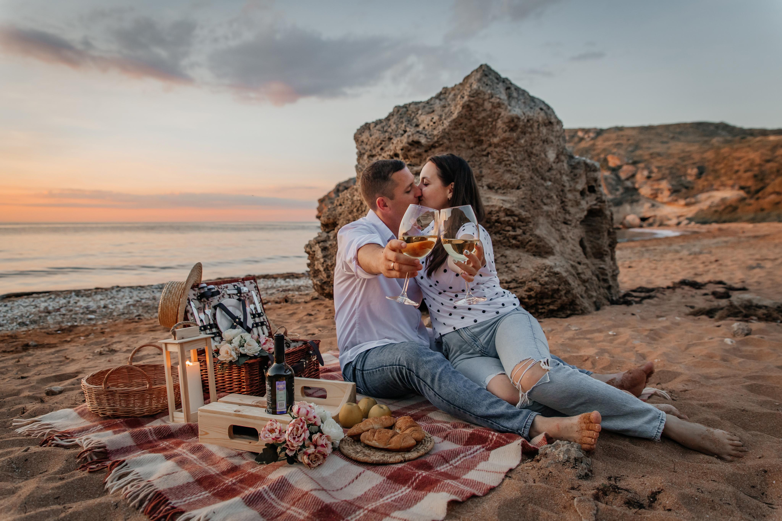 На переднем плане целующаяся пара вытянула руки вперед с бокалами вина, у них пикник сидят на пляже. 
