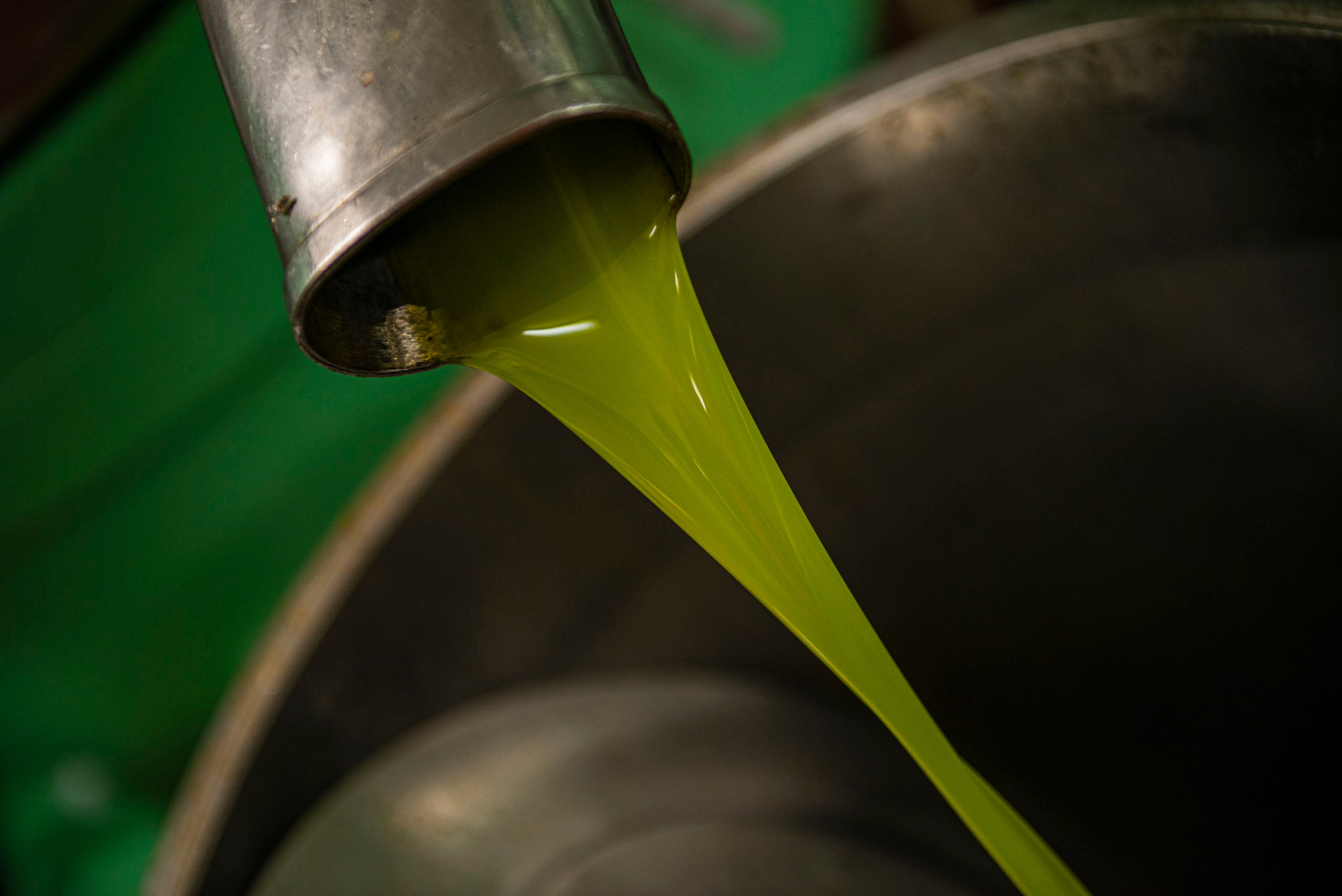 Крупный план ярко-зеленого, свежевыжатого оливкового масла extra virgin (EVOO), которое густой струей вытекает из металлического носика в сборный контейнер.