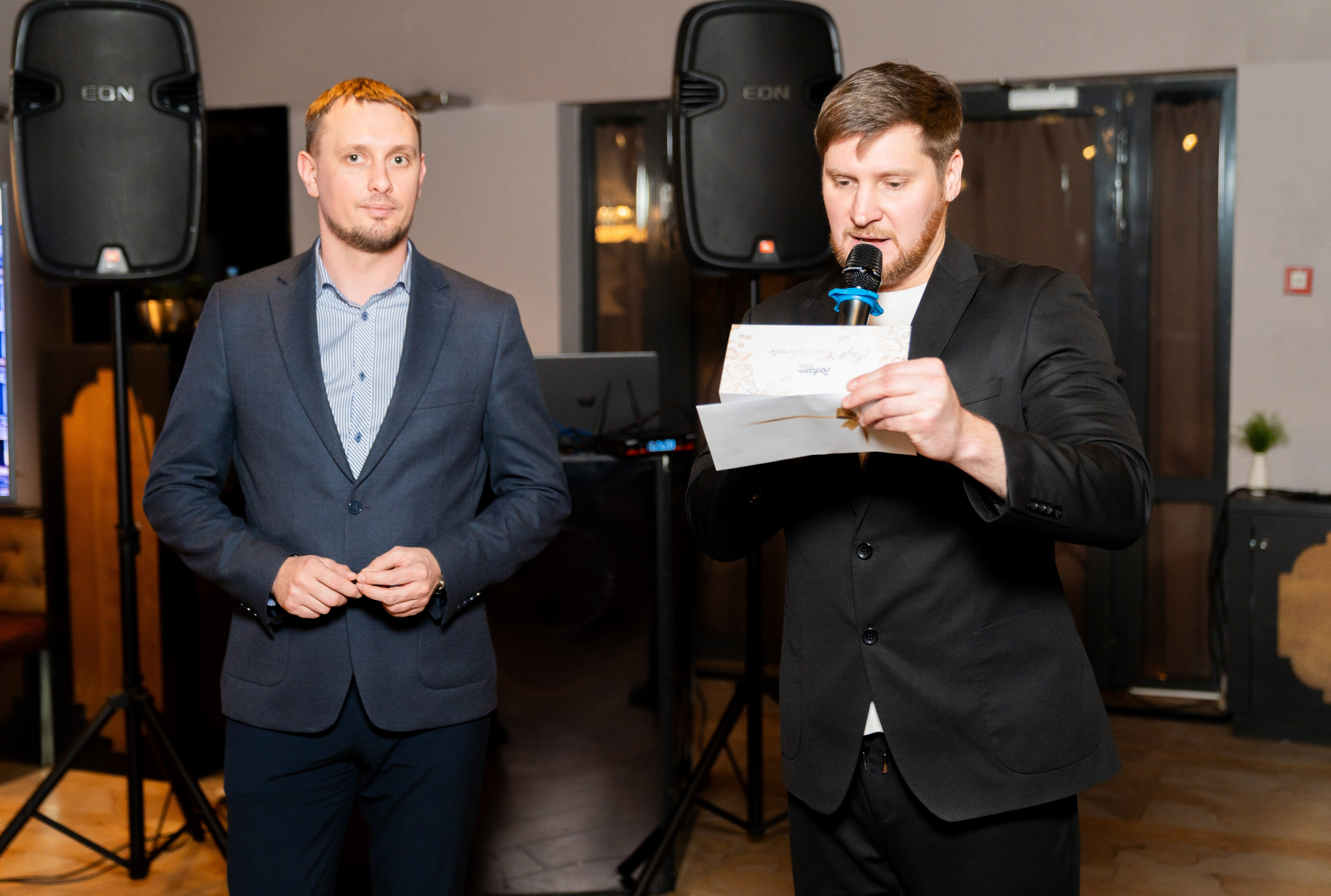 Два мужчины в костюмах на сцене: один читает подарочный сертификат Radisson, второй стоит рядом. Корпоратив Radisson Sonya. Фото Евгения Мальцева.