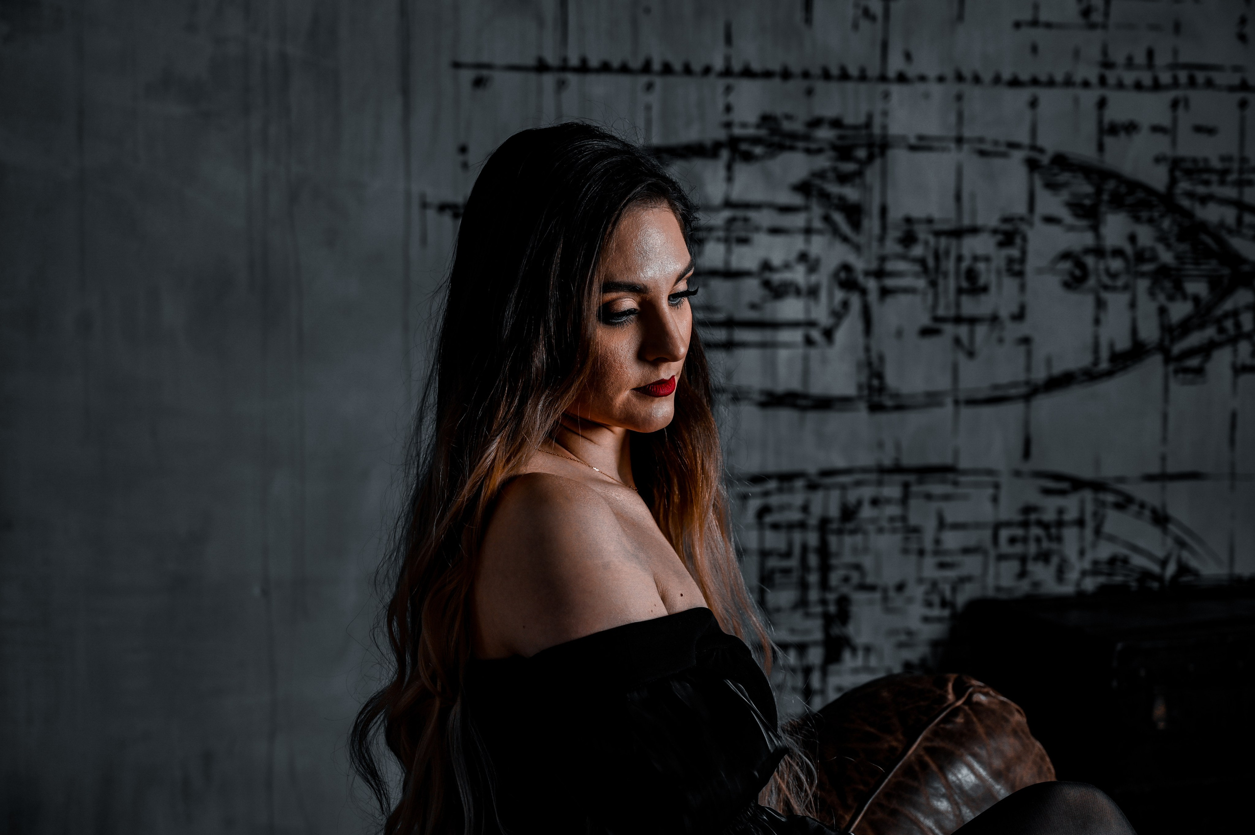 Таинственная женщина в элегантном черном платье с открытыми плечами, сидящая на стильном кожаном диване, создает атмосферу загадки и изысканности. Уникальный фон с графическими элементами подчеркивает мастерство фотографа в создании драматичных и запоминающихся портретов. Запишитесь на фотосессию сегодня, чтобы подчеркнуть свою индивидуальность!