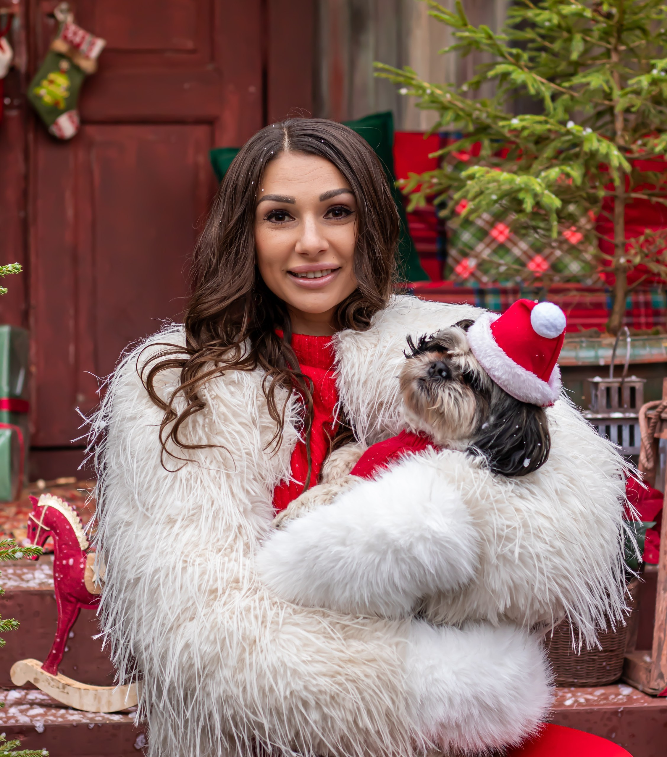 Женщина в красном с пушистой белой шубой держит собаку в новогоднем шале среди ёлок и подарков, фотограф Карасёва Елена"