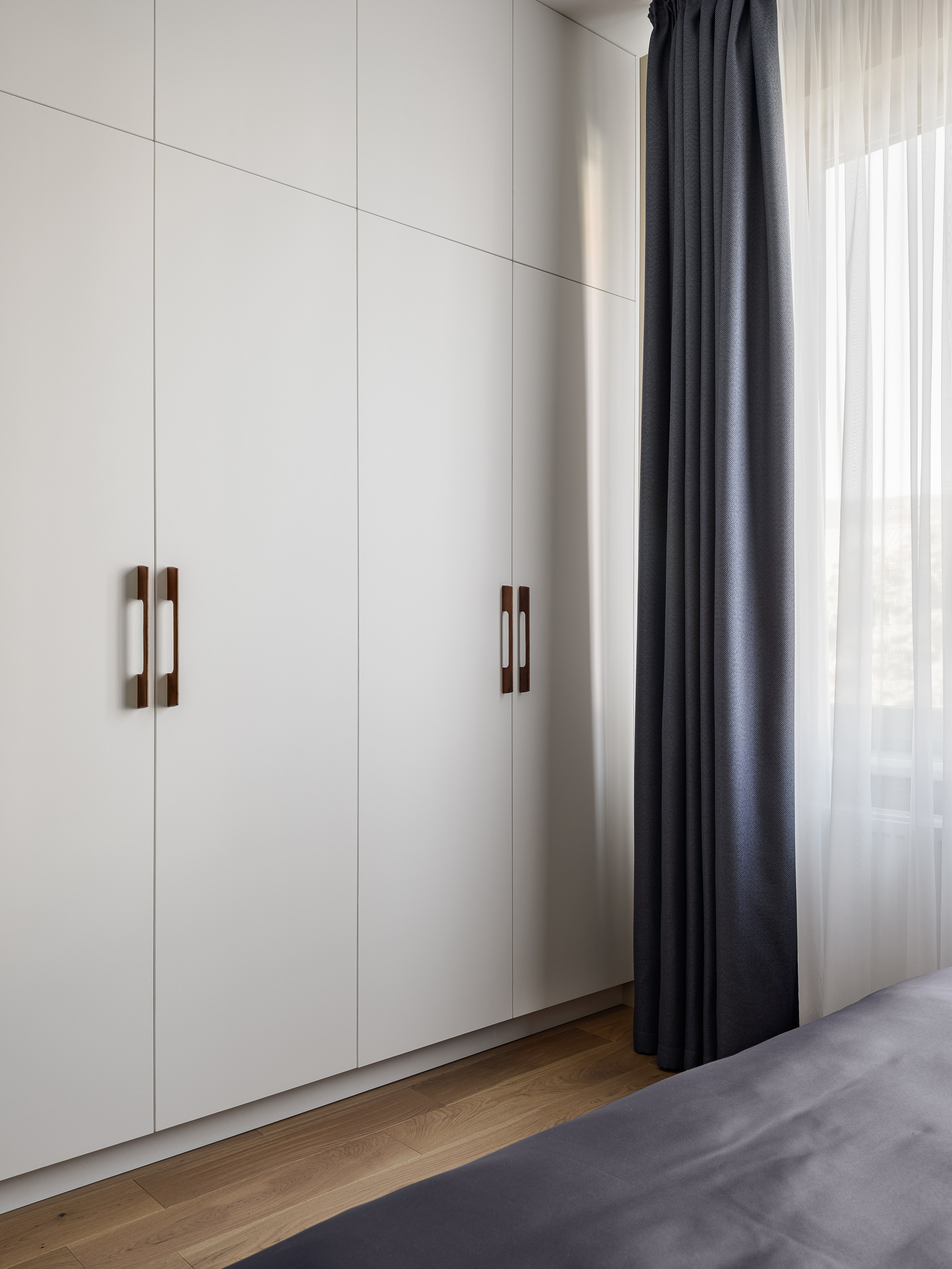 Современный встроенный шкаф с белыми фасадами и контрастными ручками в спальне, рядом длинные серые шторы и деревянный пол, дизайн Алёна Попова и Анастасия Дмитриева, фотограф Сергей Денисенко, минималистичный интерьер.