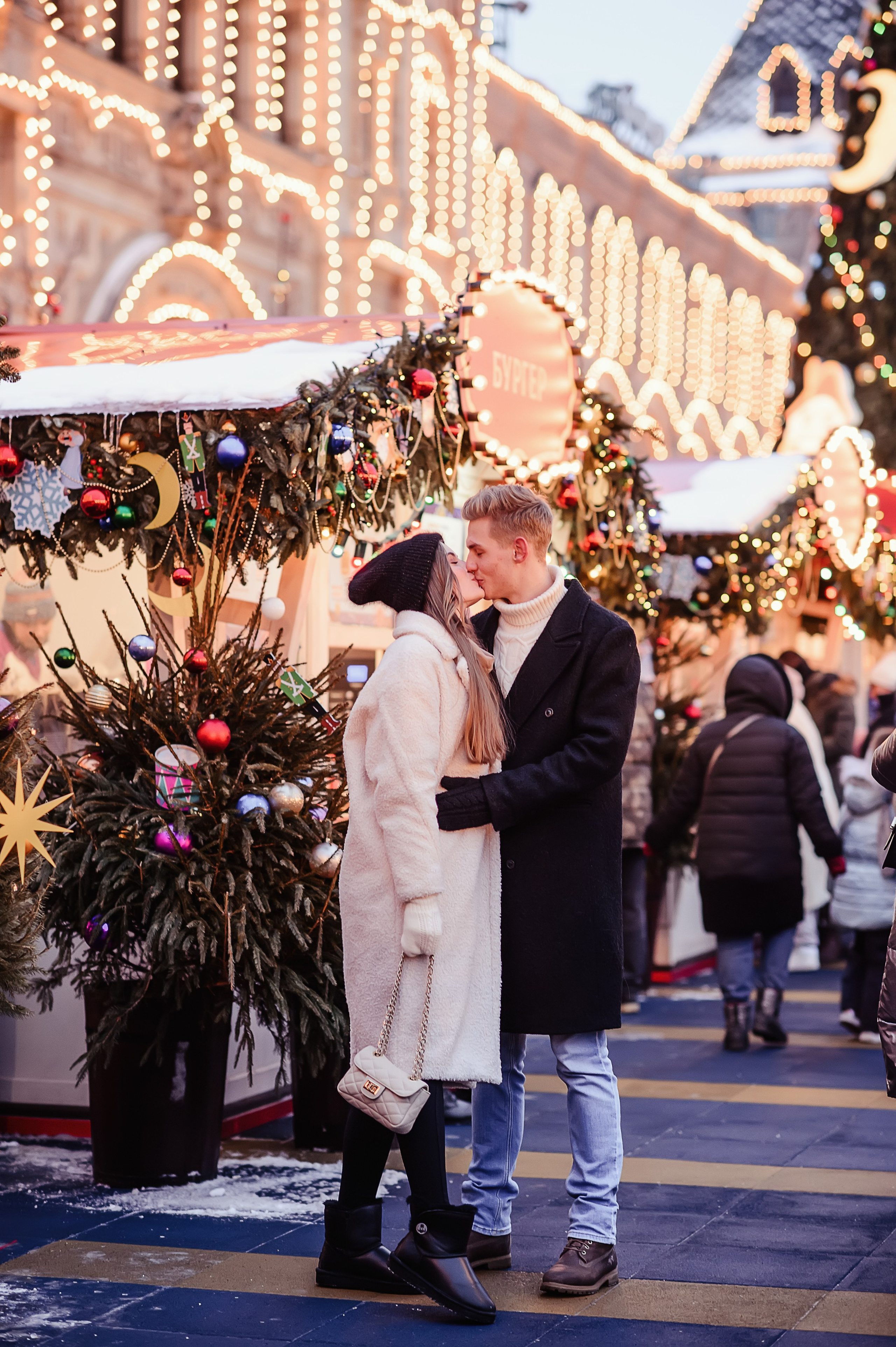 Пара влюбленных на Ярмарке на Красной площади стоят в полный рост и смотрят друг на друга на фоне огоньков ГУМа
