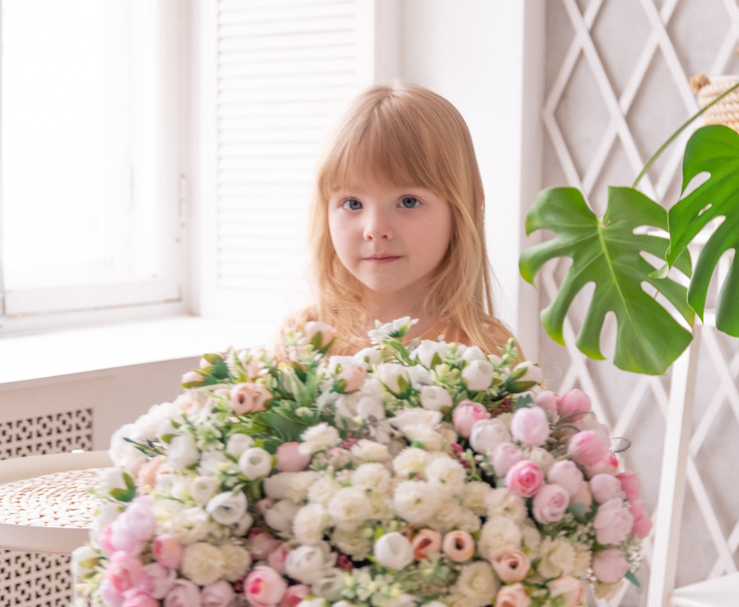 Маленькая девочка с длинными светлыми волосами держит огромный букет нежных цветов у окна — трогательный весенний портрет.
