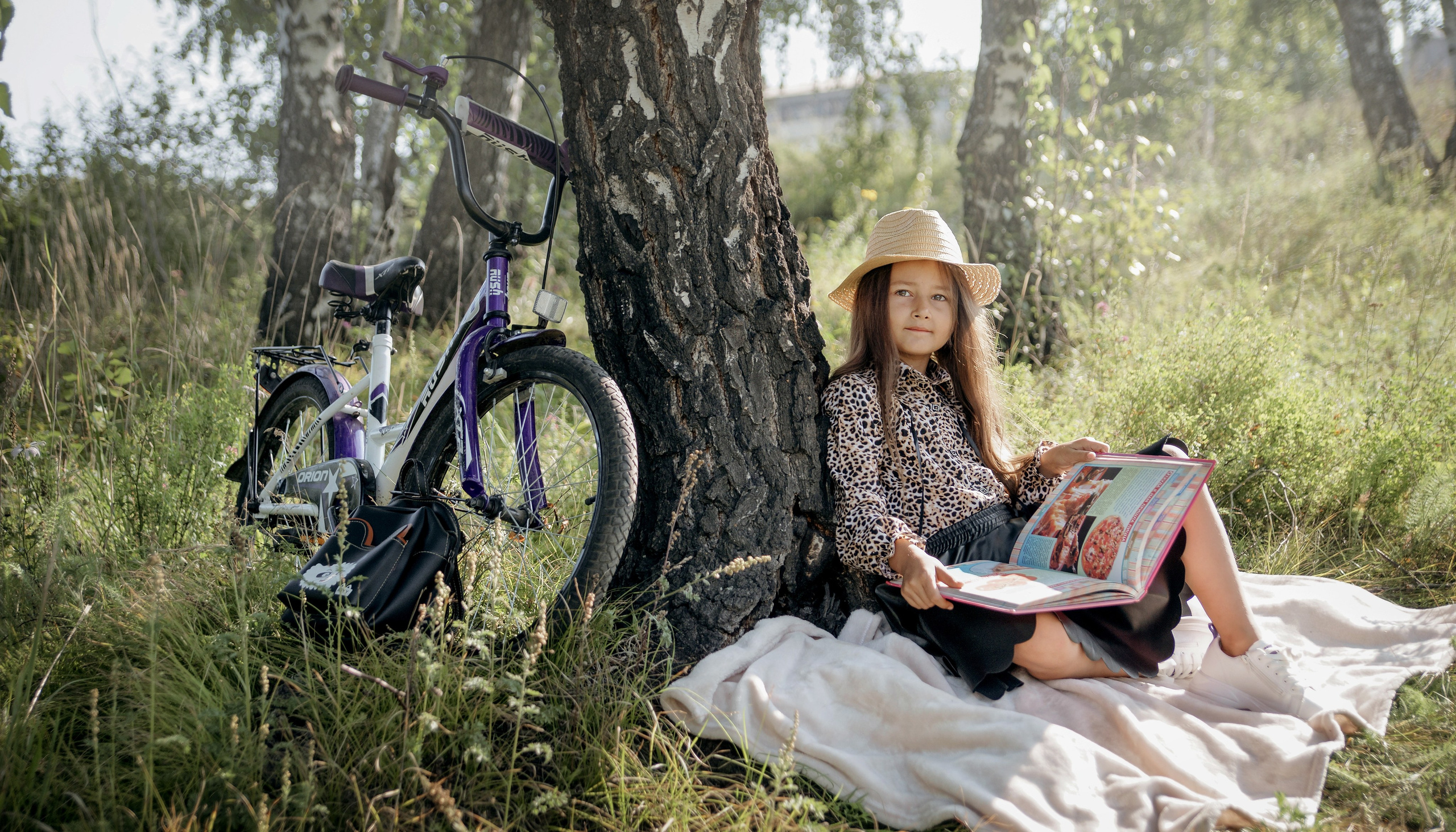 Детская фотосессия летом - девочка сидя у дерева с велосипедом и книгой
