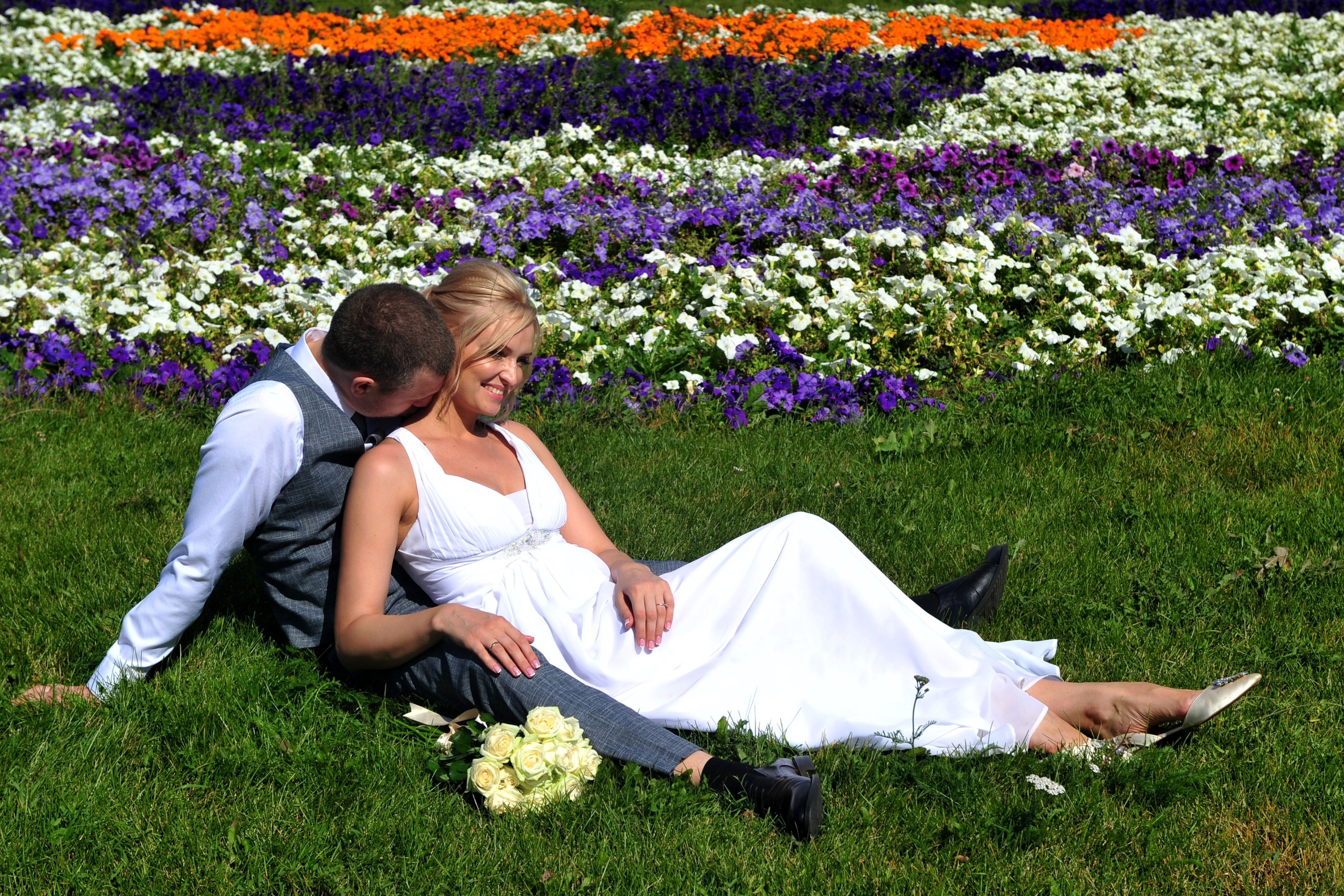 Свадьба на Воробьёвых Горах рядом с МГУ, новобрачные сидят в обнимку на зеленой траве на фоне цветочной клумбы