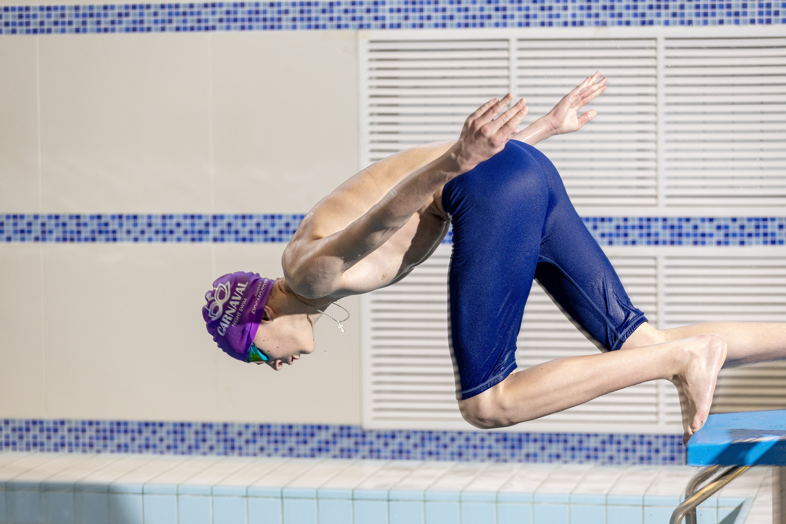 Пловец в синем купальнике и фиолетовой шапочке с надписью «CARNAVAL» совершает прыжок в бассейн с тумбы.