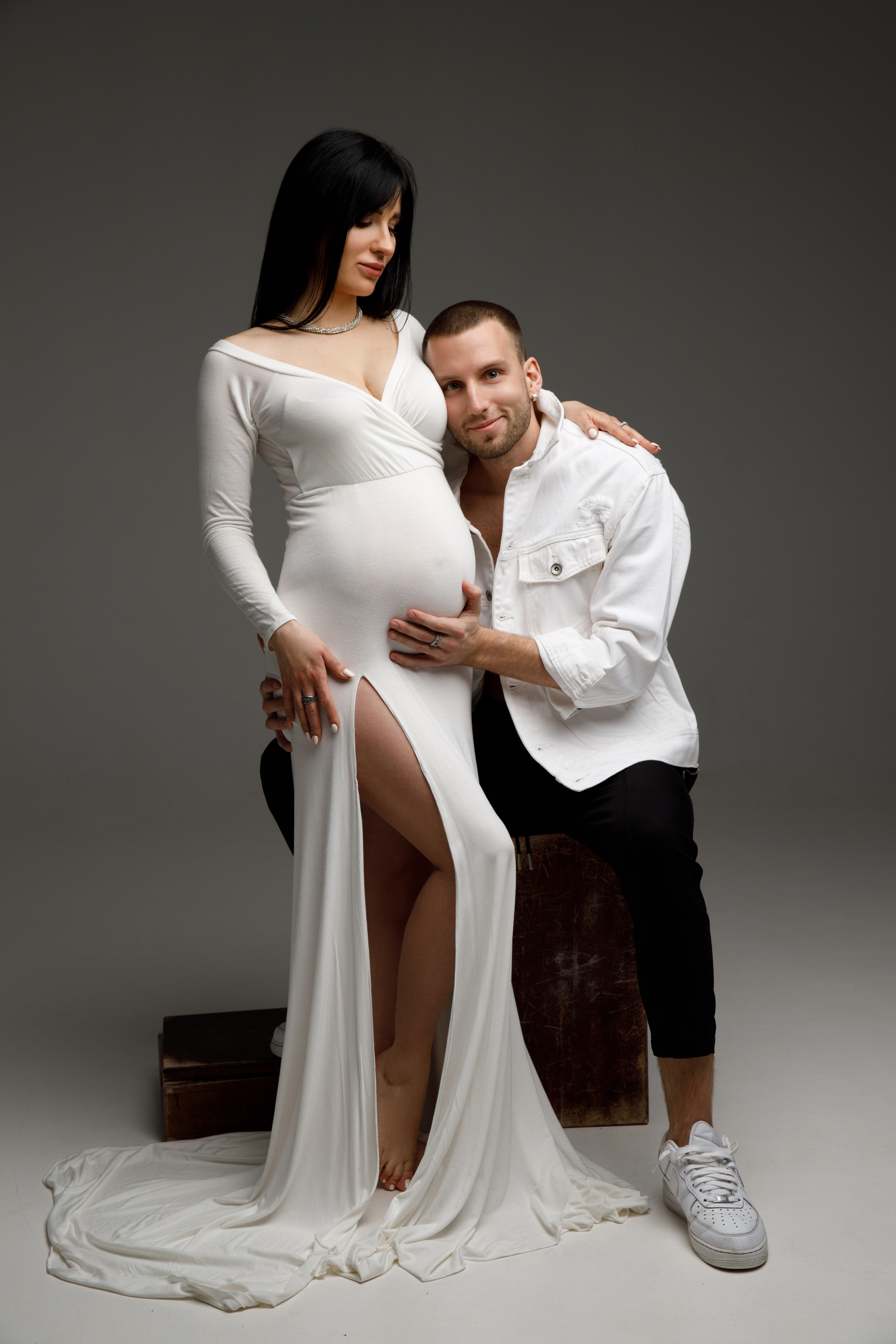 Фотосессия беременной с мужем в белом платье на сером фоне.