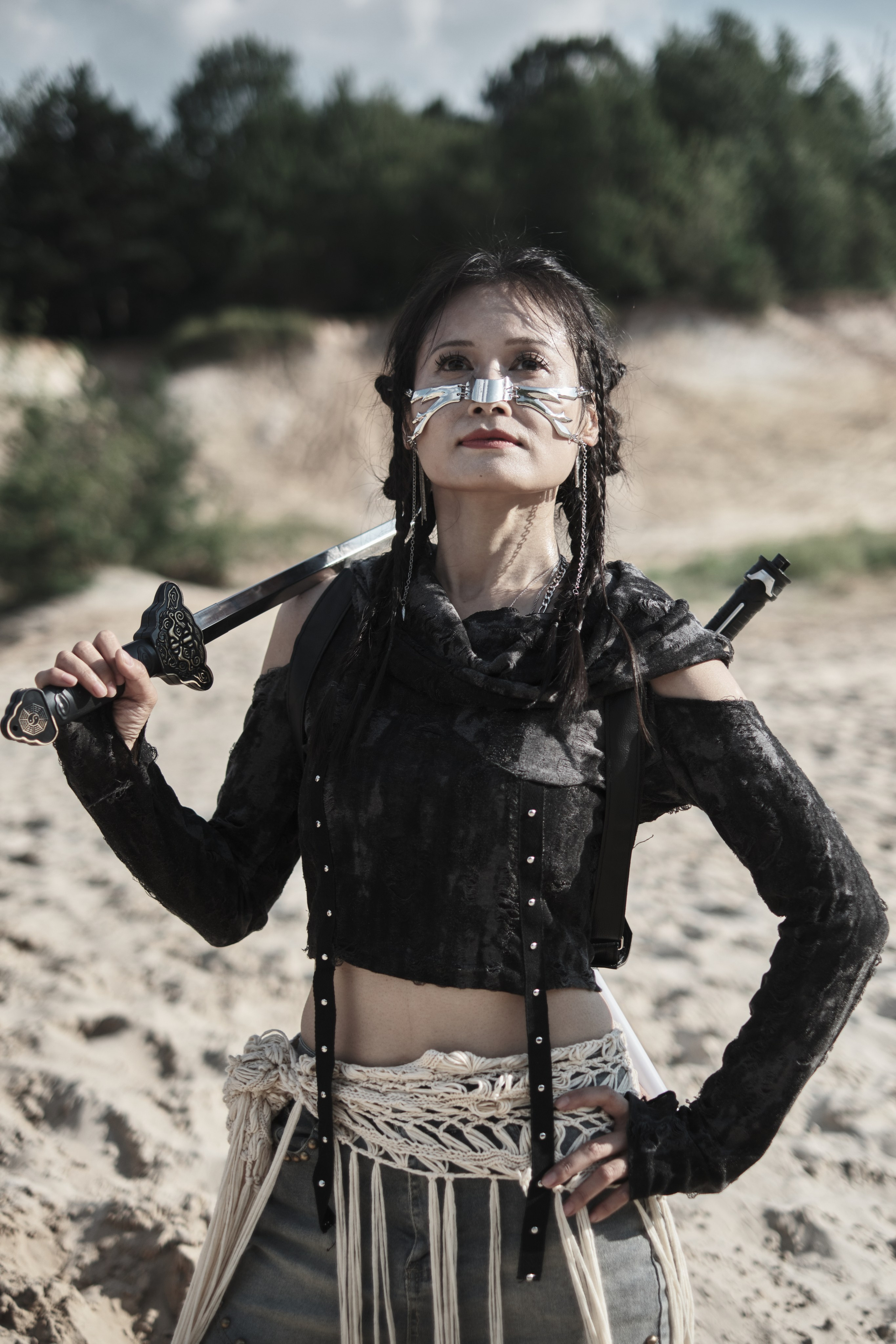 Девушка азиатской внешности и металической маской на лице стоит положив катану на спину