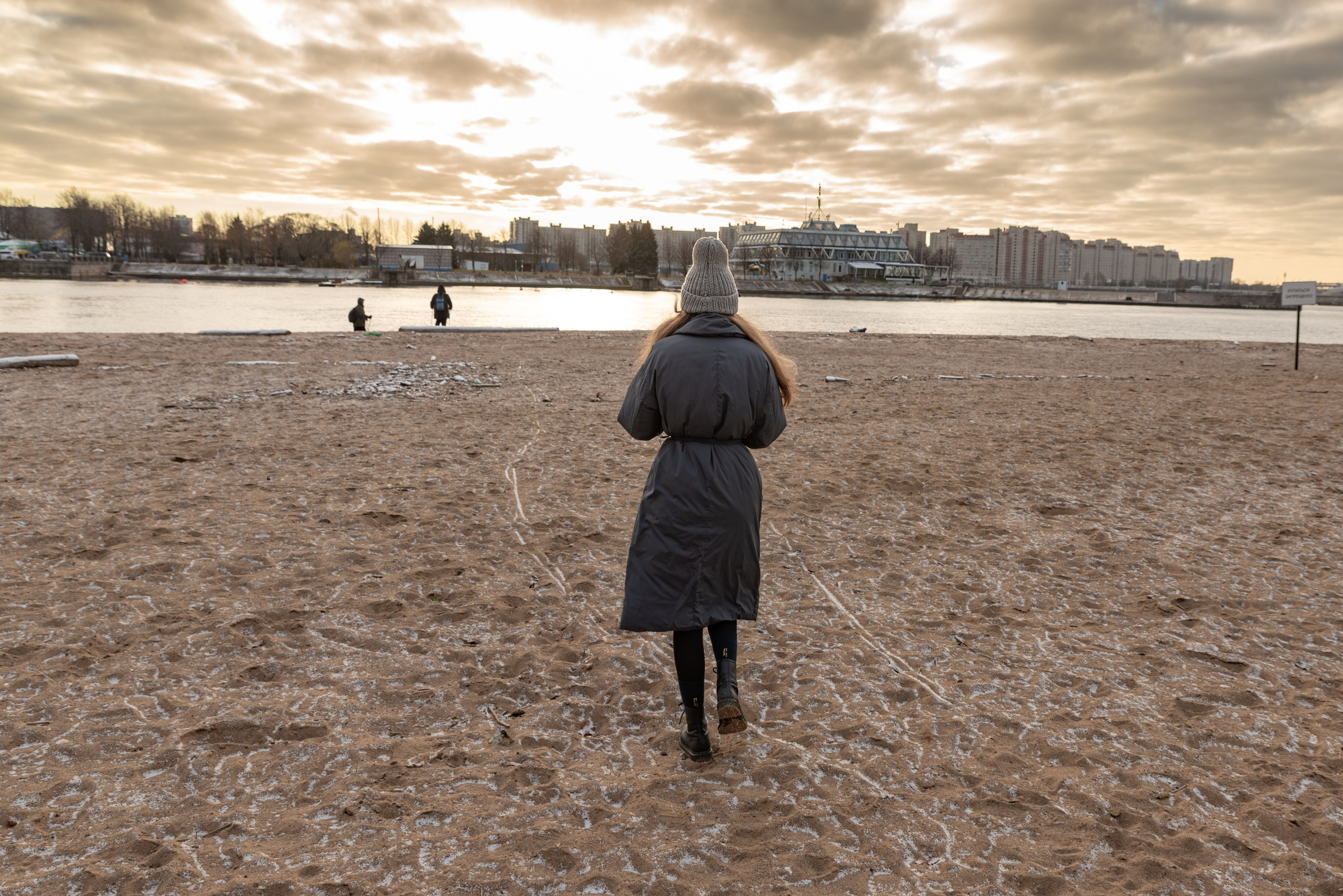 Пляж, песок, солнце светит сквозь облака. Город на другом берегу. Девушка в сером пальто идёт к воде.