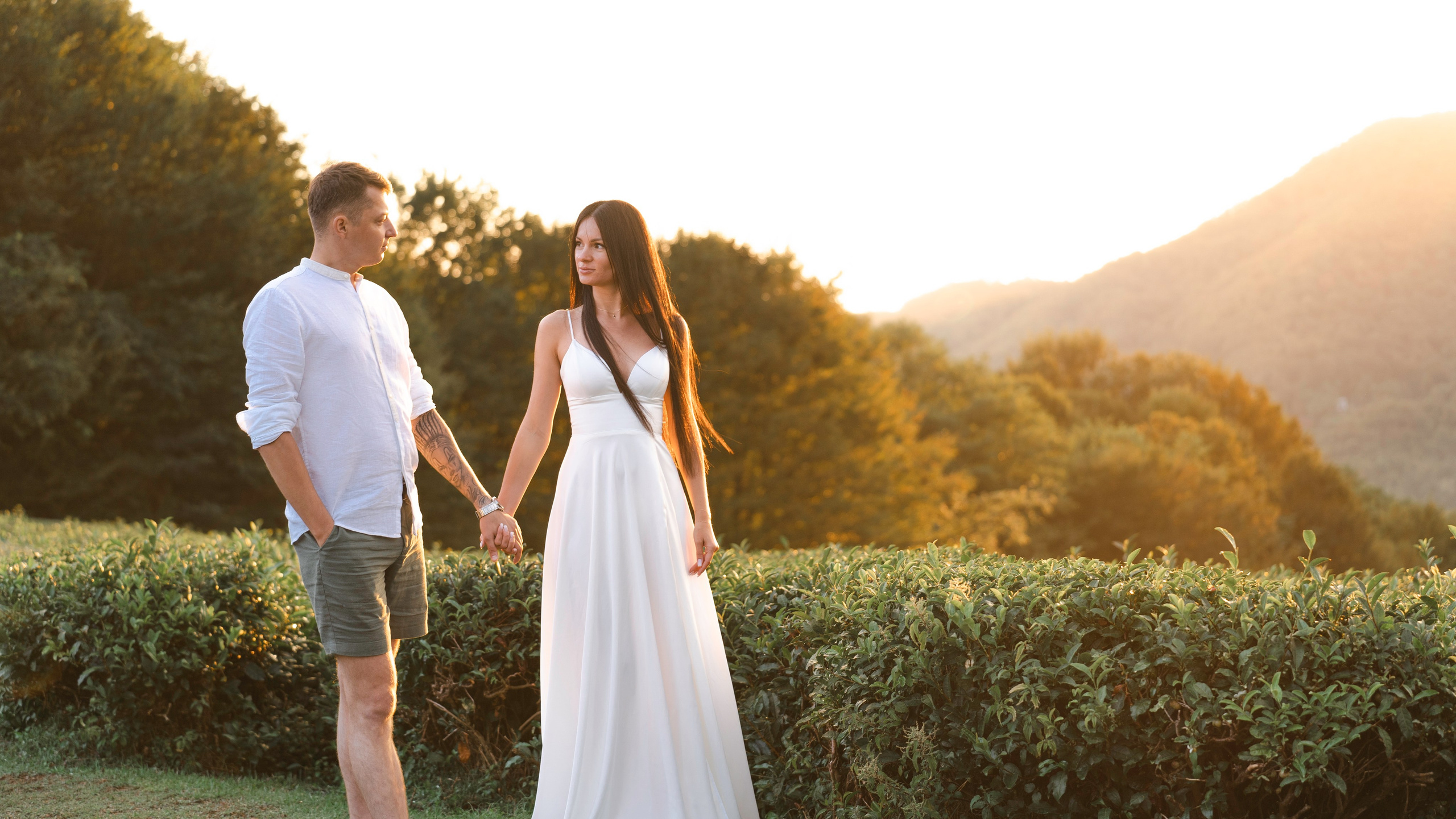 На изображении изображена пара, держащаяся за руки, на фоне живописного холмистого пейзажа, окутанного теплым сиянием заходящего солнца. Женщина одета в длинное белое платье, а мужчина - в белую рубашку и шорты. Они кажутся наслаждающимися умиротворенным моментом вместе, окруженные природой. Фотография передает атмосферу спокойствия, любви и гармонии между двумя людьми, наслаждающимися красотой окружающего мира вместе.