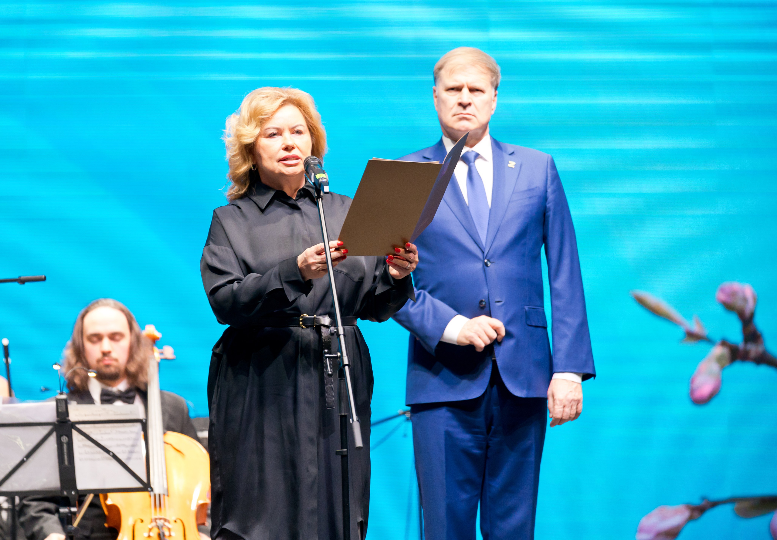 Женщина в чёрном держит папку и выступает у микрофона, мужчина в синем костюме стоит рядом. Концерт ко Дню местного самоуправления в Санкт-Петербурге. Фото Евгения Мальцев.