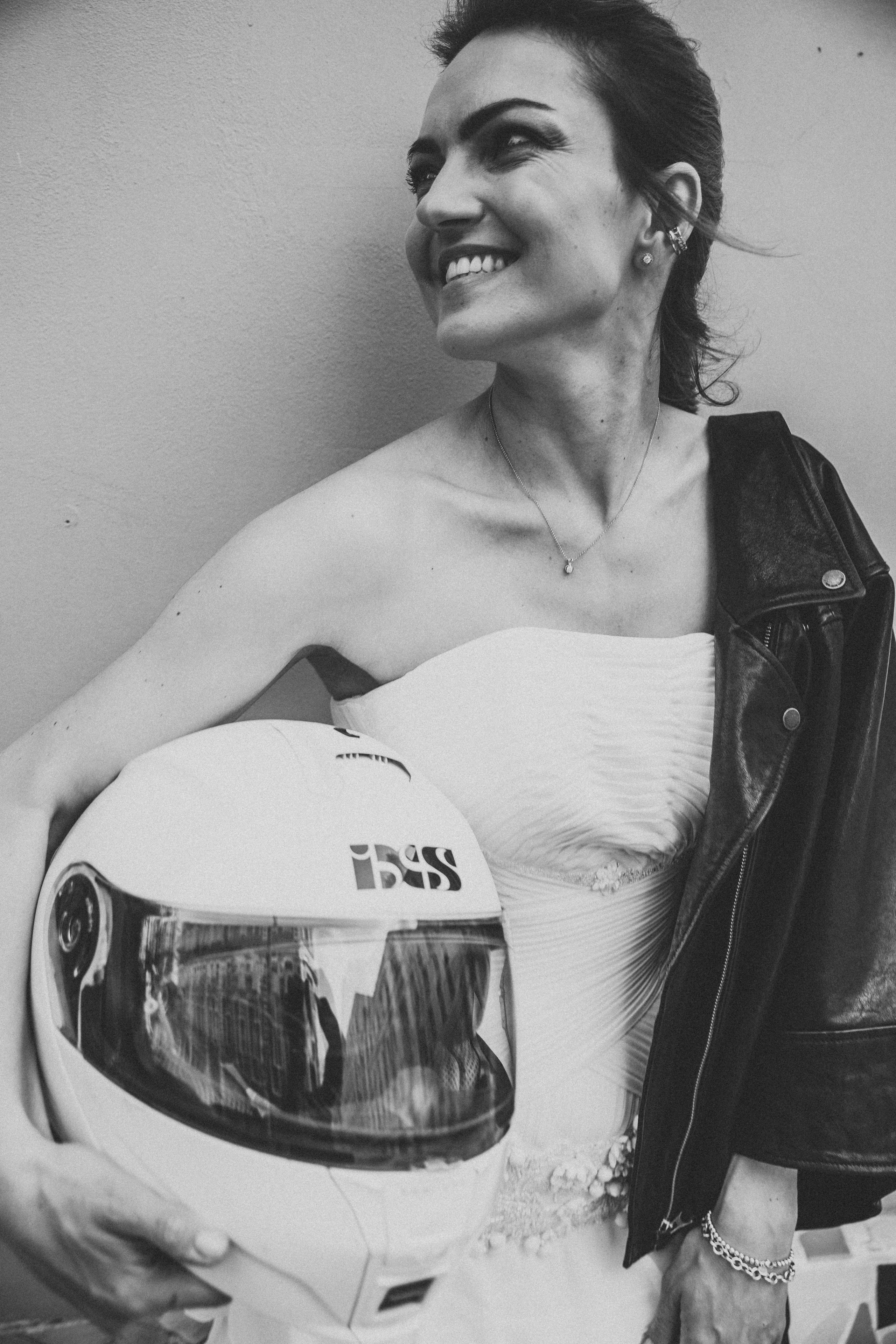 Красивая невеста держит в руках шлем от мотоцикла и улыбается своему жениху