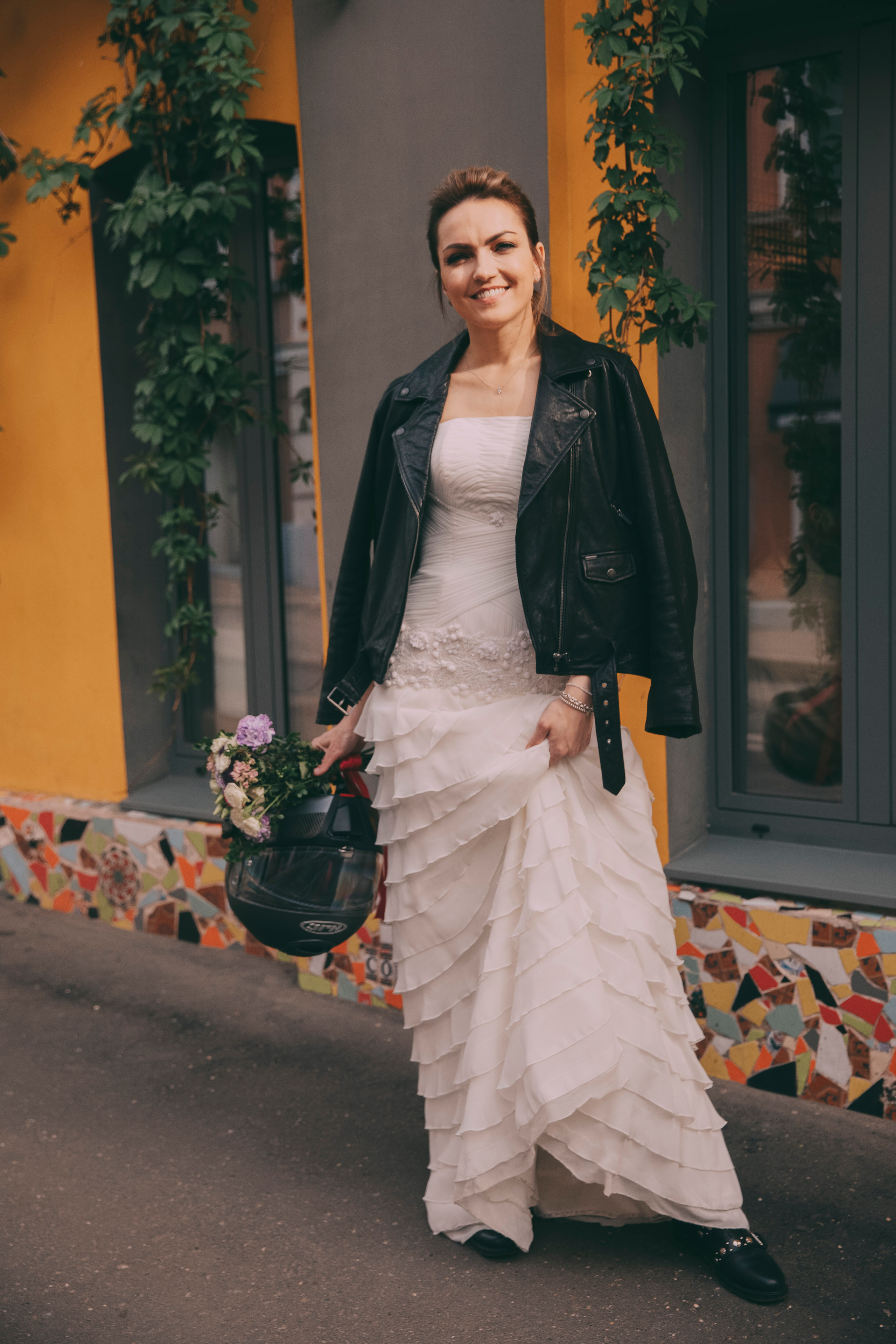 невеста в свадебном платье и кожанной куртке держит в руках шлем для мотоцикла и свой свадебный букет и улыбается своему жениху