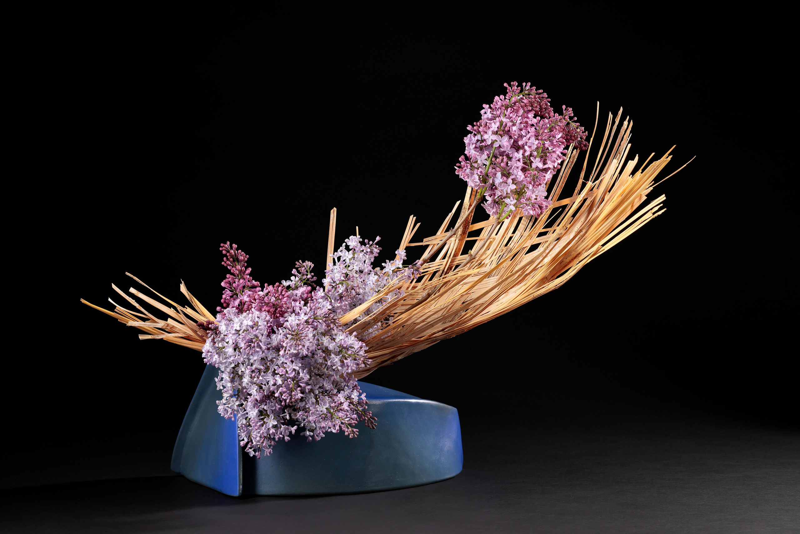 Икебана из плетёнки и сирени в голубой дизайнерской вазе на чёрном фоне.