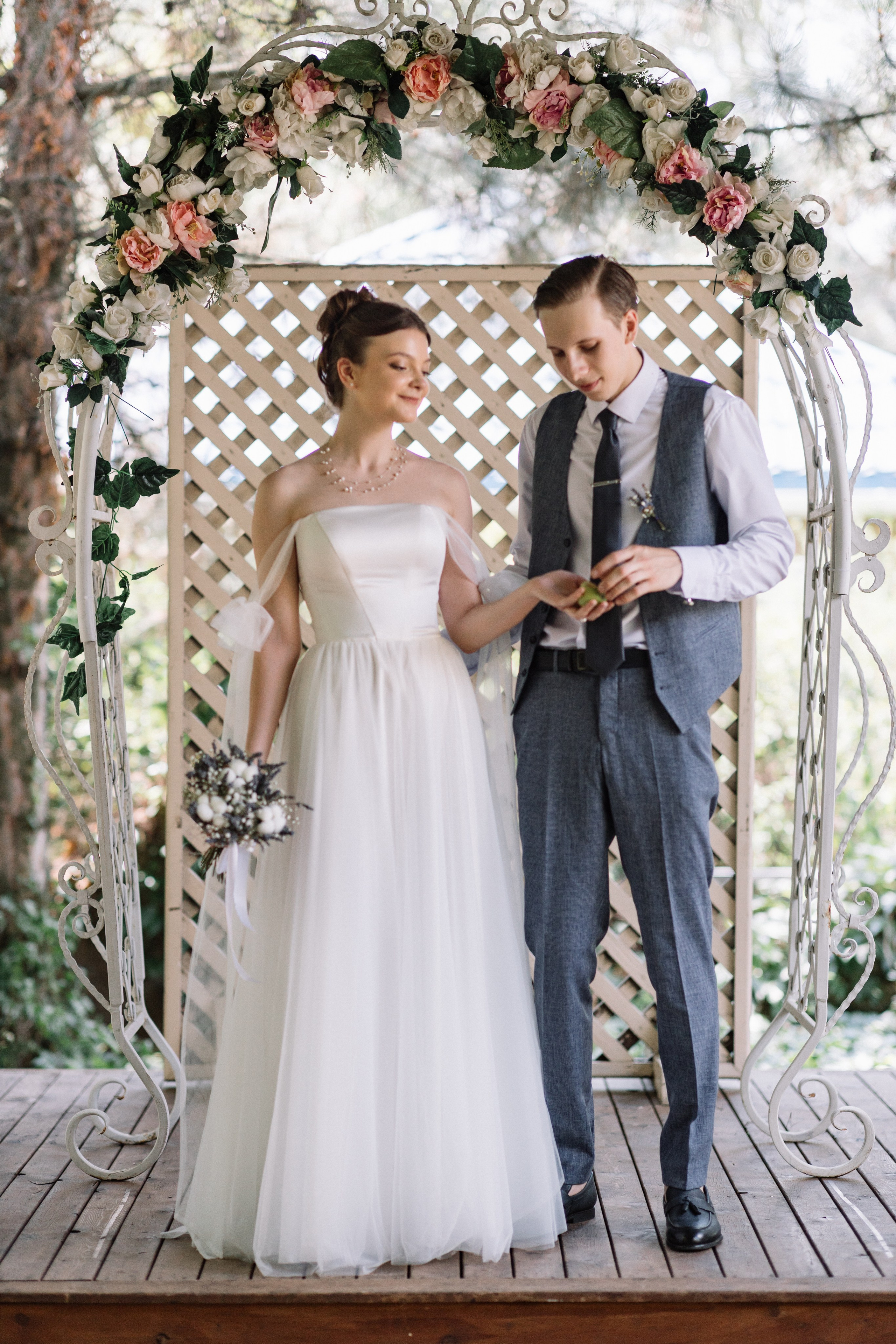 Молодожены целуются под аркой из цветов, белые ленты и зелень вокруг.