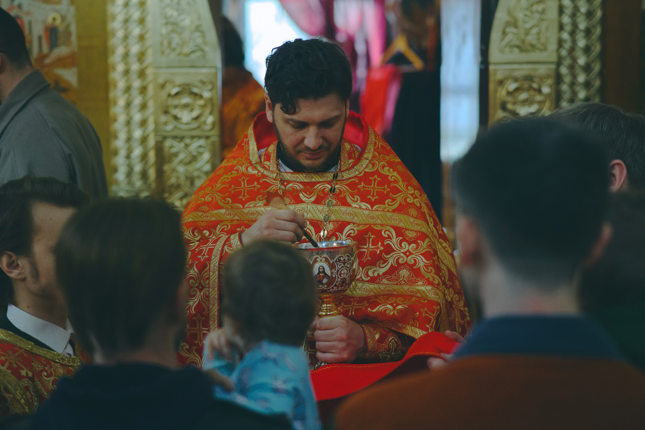 Священник в красных облачениях держит евхаристическую чашу и ложицу, совершая причастие. В первом ряду — прихожане, включая ребёнка.