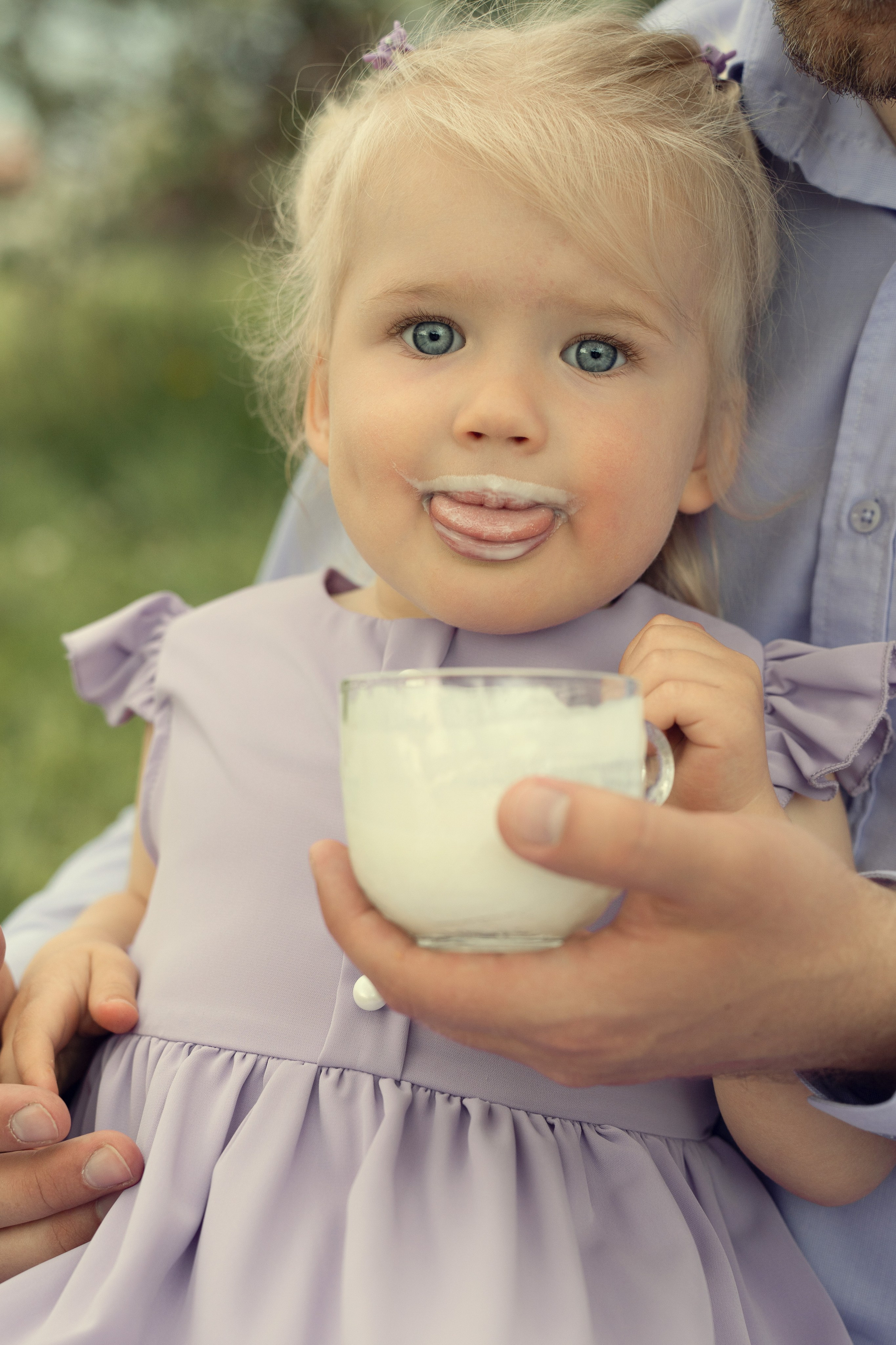 Маленькая девочка смешно пьет кефир из кружки в парке.