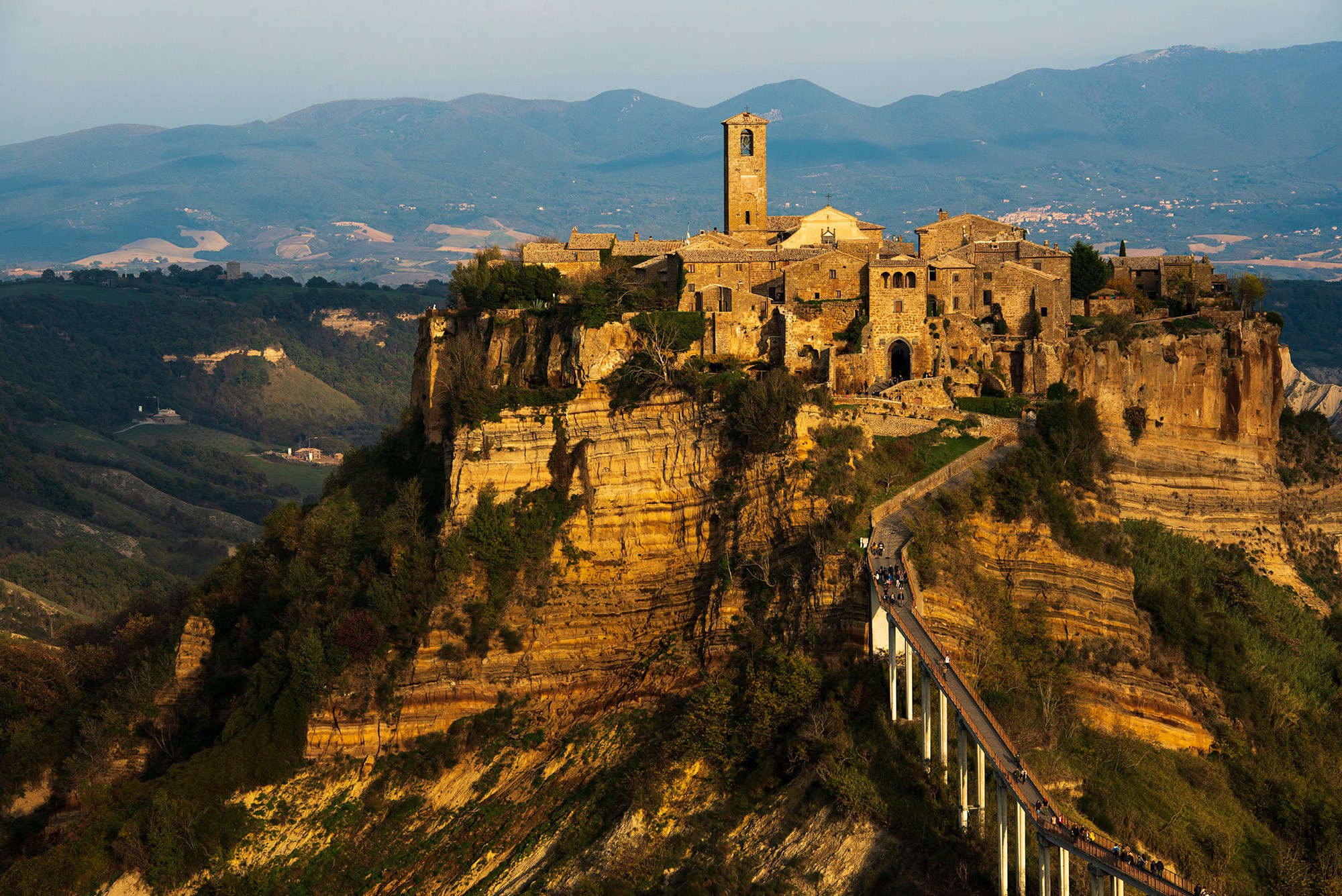 Художественный пейзаж 'умирающего города' Чивита-ди-Баньореджо в Италии: панорамный вид средневековой деревни на вершине туфовой скалы, длинный пешеходный мост, соединяющий с материком, мягкое закатное освещение.
