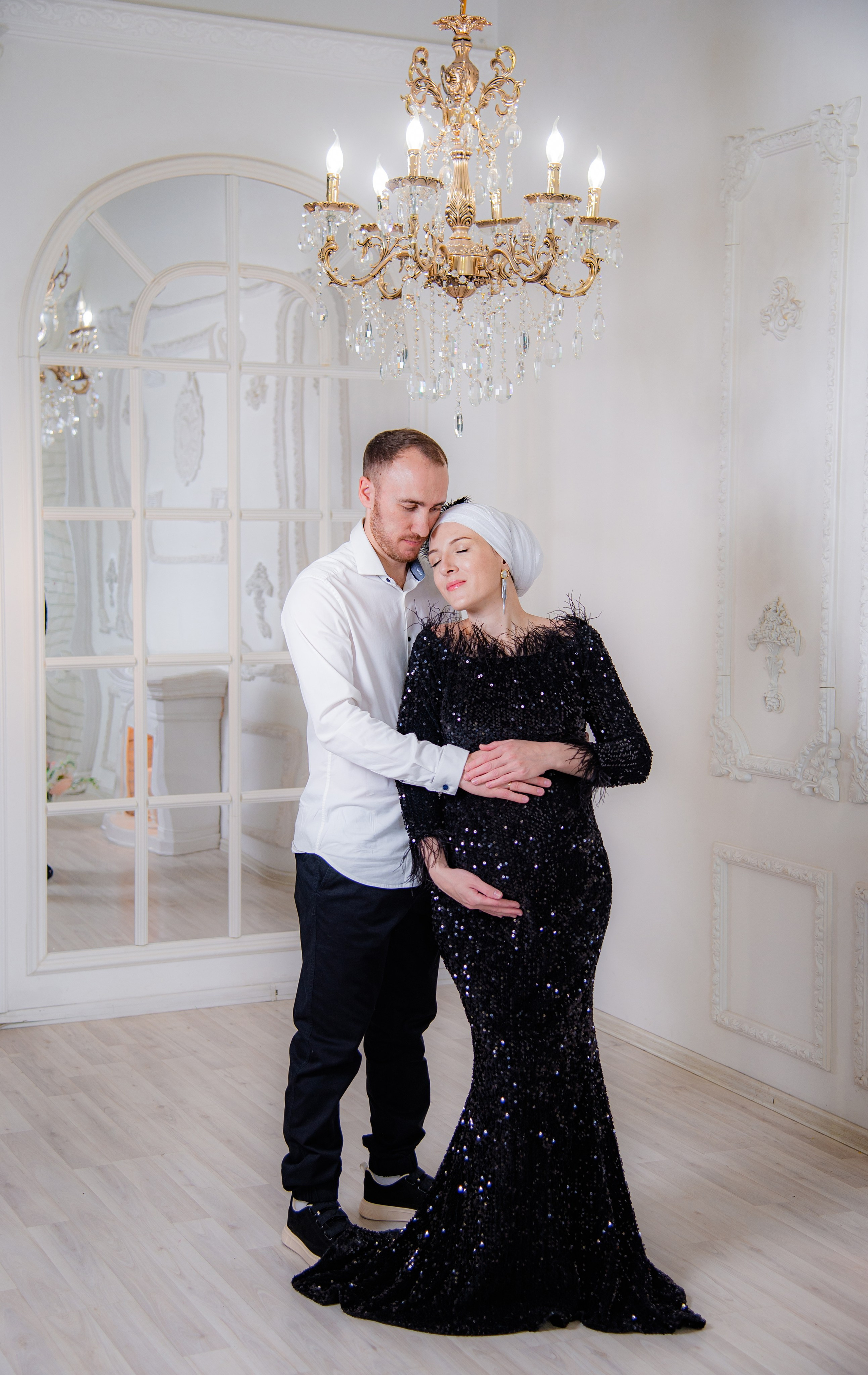 Фотосессия беременных в фотостудии в Уфе: будущая мама в элегантном платье из проката, с профессиональным макияжем и прической. Она счастливо позирует на фоне стильного интерьера, а рядом ее муж и члены семьи. Фотограф помогает в позировании, создавая теплую и уютную атмосферу, отражающую красоту и нежность материнства