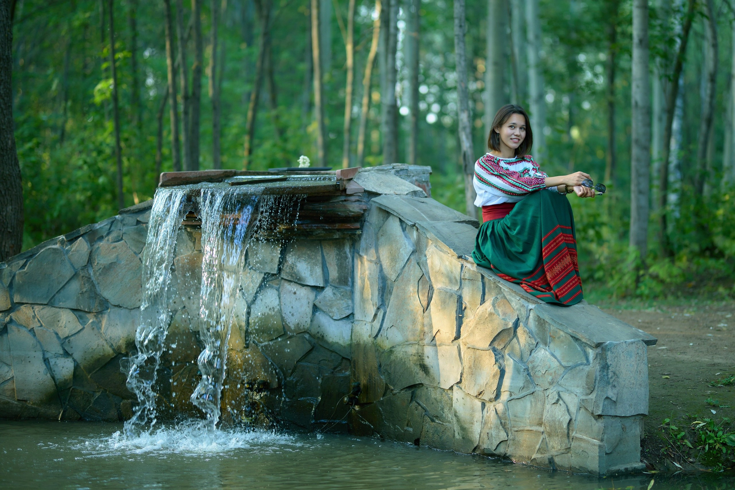Девушка в национальном костюме сидит в парке около пруда