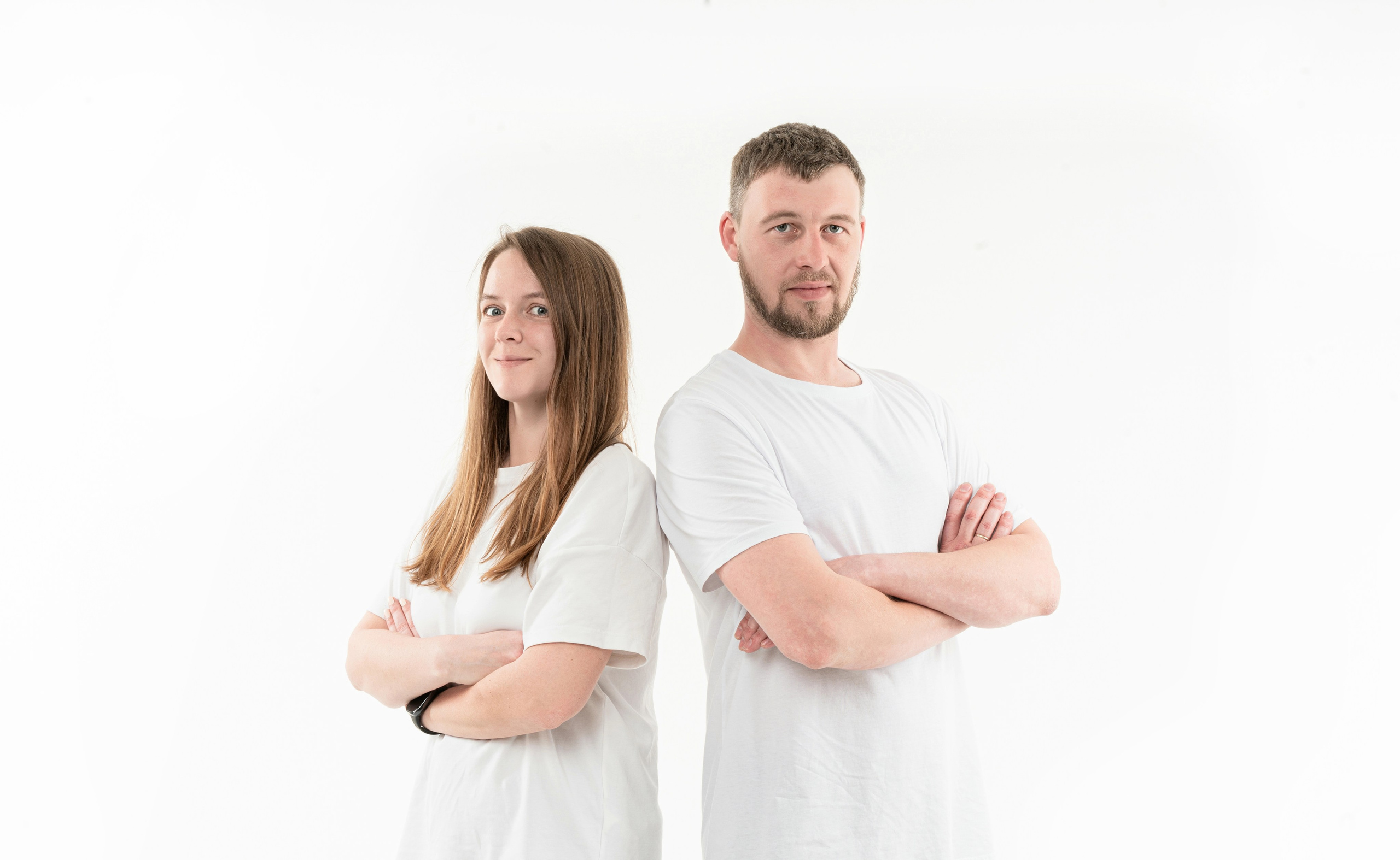 На изображении изображены два человека: мужчина и женщина, стоящие спиной к спине с перекрещенными руками. Оба одеты в простые белые футболки и кажутся уверенными или серьезными. Фон ярко-белый, что делает основные объекты визуально выделенными. На изображении отсутствует какой-либо текст.