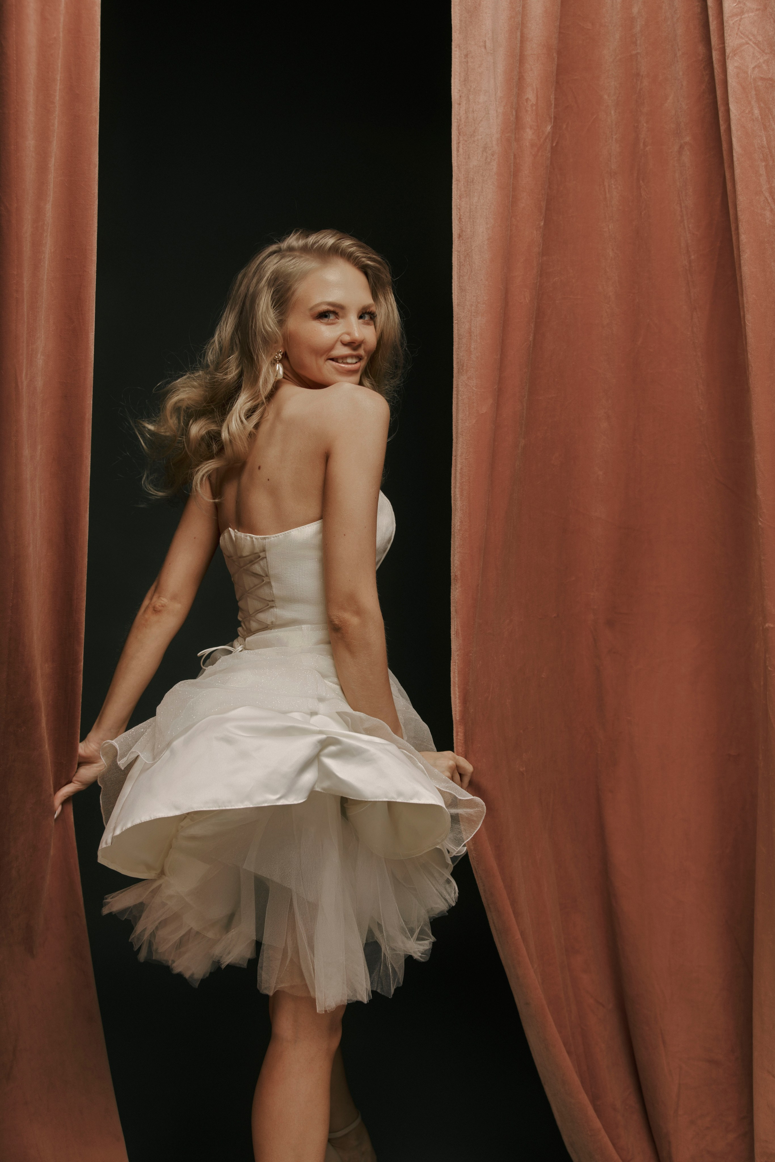 Две женщины в вечерних платьях на фоне гардеробной стойки с одеждой. Одна из них стоит в ярко-красном платье с объемным декором в виде роз, а другая сидит в воздушном белом платье. Интерьер оформлен в темных тонах с элементами драпировки. На переднем плане заметны книги и туфли на высоких каблуках.