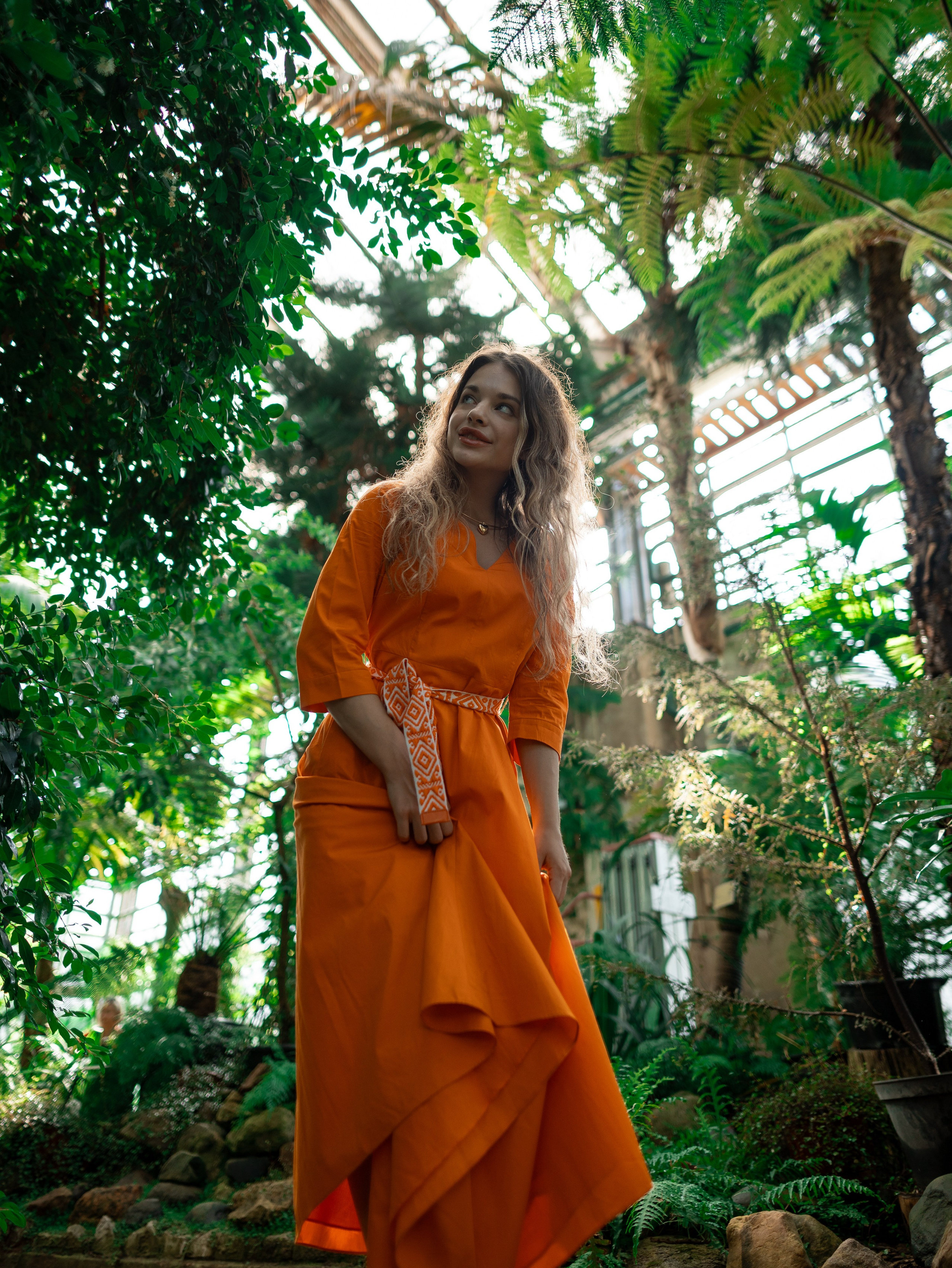 Девушка в оранжевом платье стоит на фоне зелени