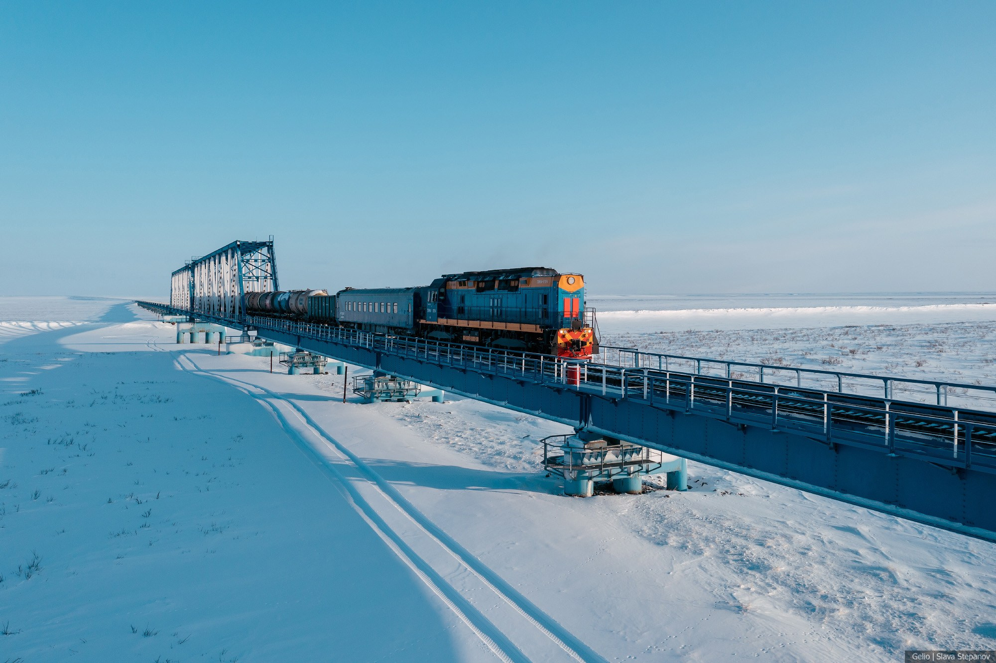 Локомотив с вагоном на ажурном мосту через заснеженную тундру, аэрофотосъёмка самой северной железной дороги в лучах низкого зимнего солнца, фото Slava Stepanov.