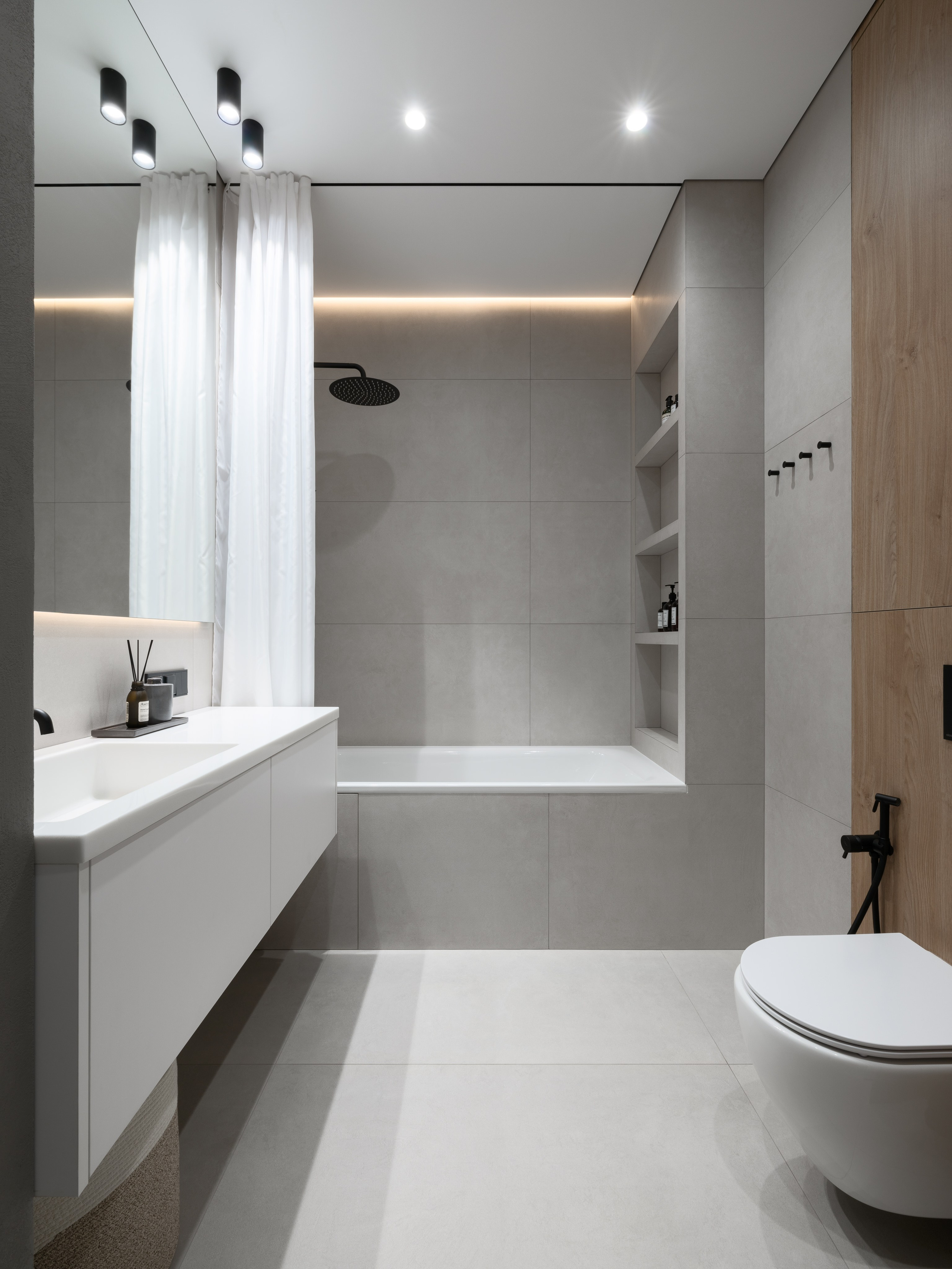 Современная ванная комната в минималистичном стиле с подвесным унитазом, белым умывальником и ванной с душем. В отделке серый керамогранит и панели под дерево. Автор проекта — дизайнеры Евгения Княжева и Владлена Привалова, фотограф Сергей Денисенко